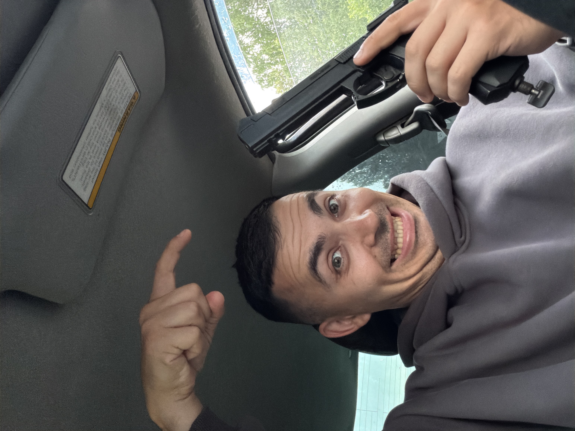 A man is taking a selfie inside a car. He is wearing a grey hooded sweatshirt and has short dark hair. He is making a surprised expression with his eyes wide open and mouth agape. He is pointing one finger up with his left hand, and in his right hand, he is holding a black pistol. The pistol appears to be a toy or replica.
The setting is inside a car, with the sun visor visible on the left side of the frame. The visor has text on it, which appears to be a warning label. The window behind the man shows trees and greenery, suggesting it is daytime and possibly outdoors. The lighting is bright, indicating daylight. The man's expression and the object in his hand might suggest a playful or provocative gesture, but without further context, it is difficult to determine the exact sentiment. The cropped images highlight the man's face, the pistol, his hands, and the sun visor with its warning. The text on the sun visor is partially visible and reads "A WARNING DEATH OR SERIOUS INJURY can occur".