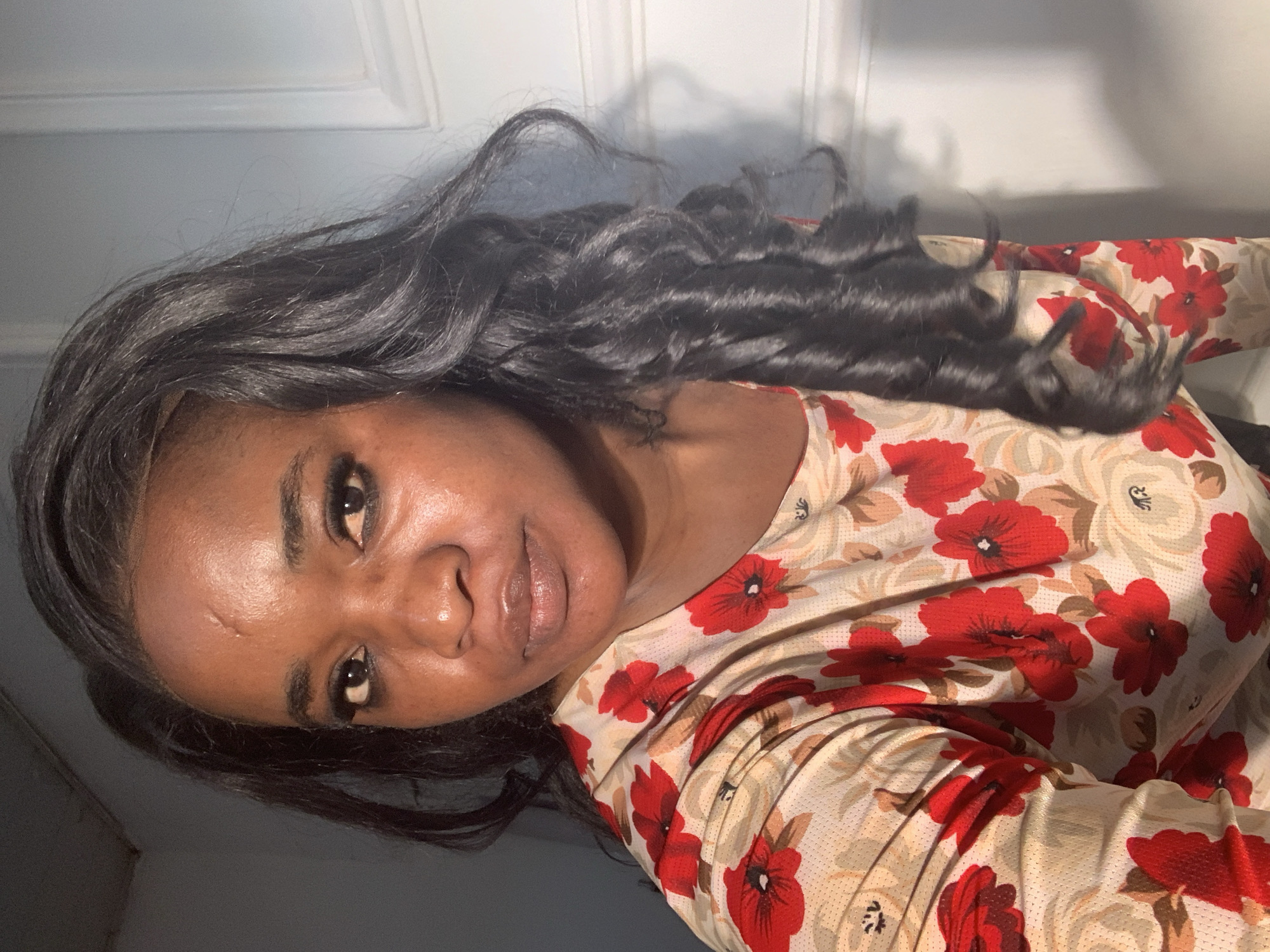 The image shows a person with dark skin and long, dark, curly hair. They are wearing a floral-patterned shirt with red and beige flowers. The background appears to be a white wall. There is no indication of the location being Barakin Akawo, Nigeria. No other individuals or objects are visible. The person is looking directly at the camera.