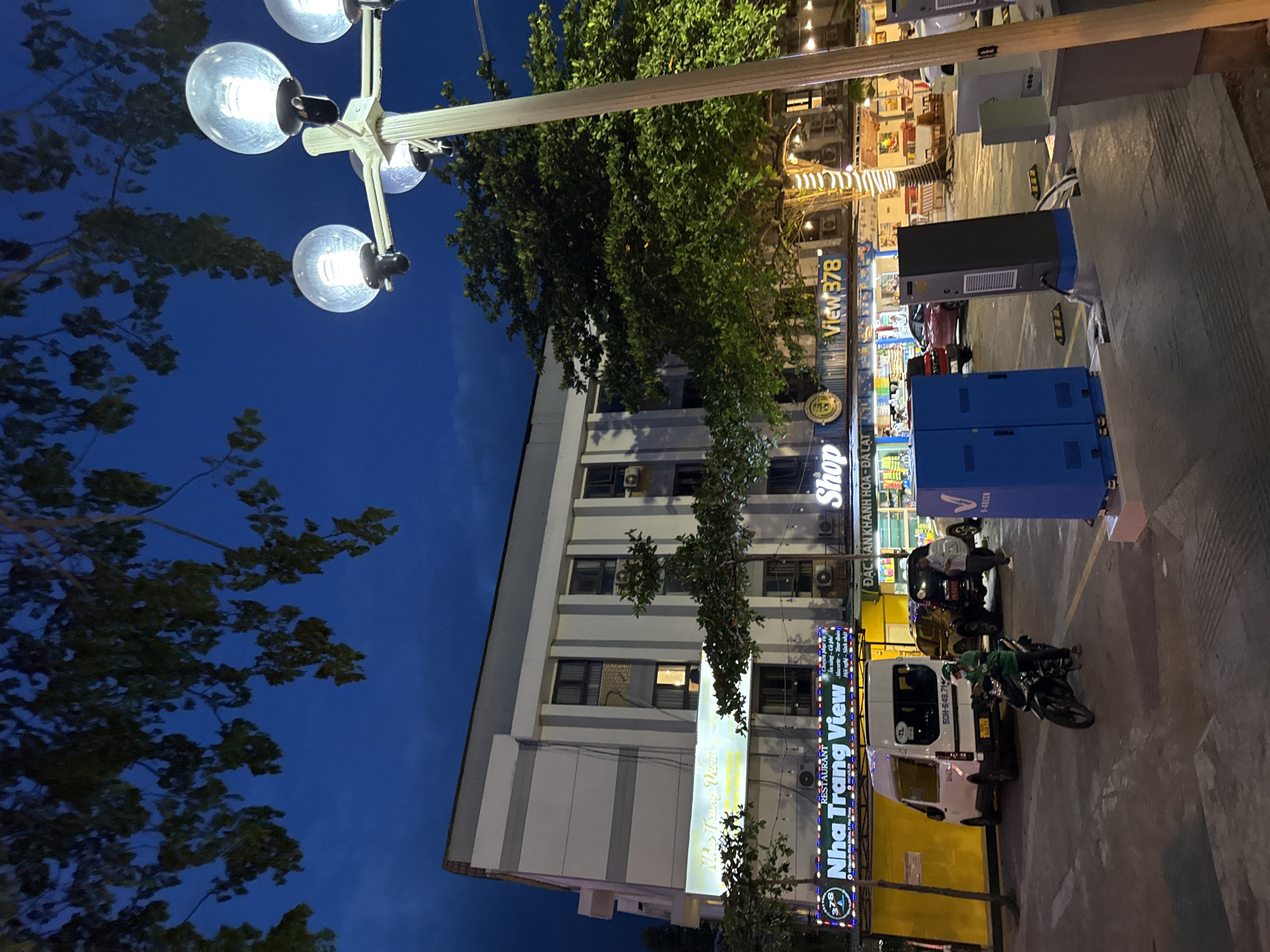 The image depicts a street scene at dusk in Nha Trang, Vietnam, specifically in front of the "Nha Trang View" establishment, a restaurant and hotel complex. A multi-story building with a light-colored facade and horizontal grey bands dominates the background. The building features numerous windows and air conditioning units. In the foreground, a paved area appears to be a parking lot.
Several vehicles are present: a white van with the license plate "50H-648.71" parked on the left, a black SUV behind it, and a red car further to the right. A person on a dark-colored motorcycle, wearing a green uniform and helmet, is in the process of riding through the parking lot. Two men are standing near the black SUV, one appearing to converse with the other.
In front of the "Nha Trang View" building, illuminated signs indicate a restaurant and shop. One sign reads "RESTAURANT Nha Trang VIEW" in prominent lettering, accompanied by smaller text. Another sign displays "Shop" and "View378". Yellow and blue rectangular charging stations or electrical boxes are visible in the foreground.
A lamppost with multiple spherical lights stands to the right of the frame, illuminating the scene. Overhanging tree branches with green leaves are visible in the upper portion of the image, obscuring the deep blue twilight sky. The overall atmosphere is that of a commercial area in a coastal city during the early evening hours.