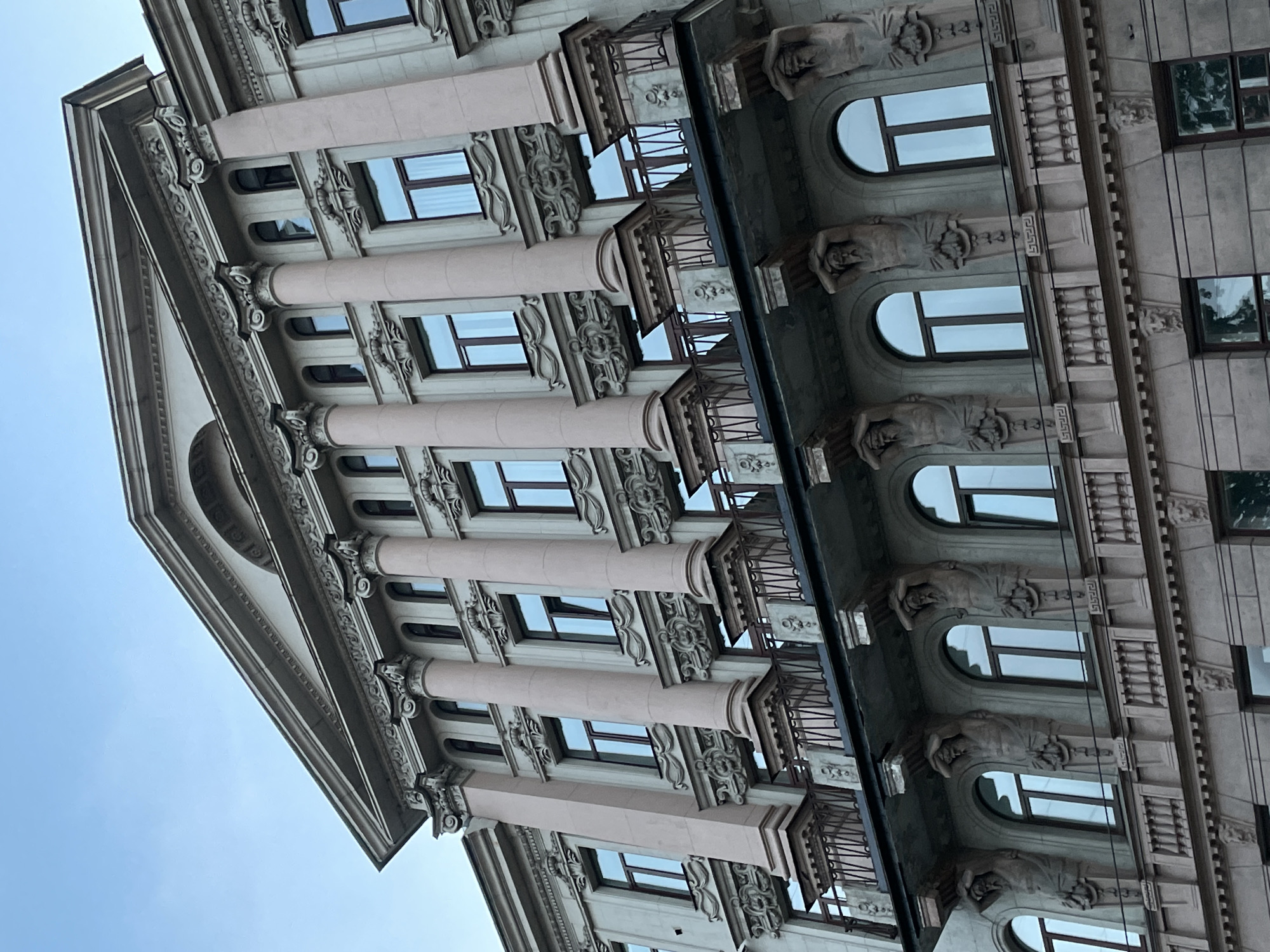 The image shows the facade of a building in Saint Petersburg, Russia. The building features classical architectural elements, including columns, ornate window frames, and decorative statues of human figures. The building has a combination of rectangular and arched windows, as well as balconies with wrought iron railings. The sky is visible in the background.
