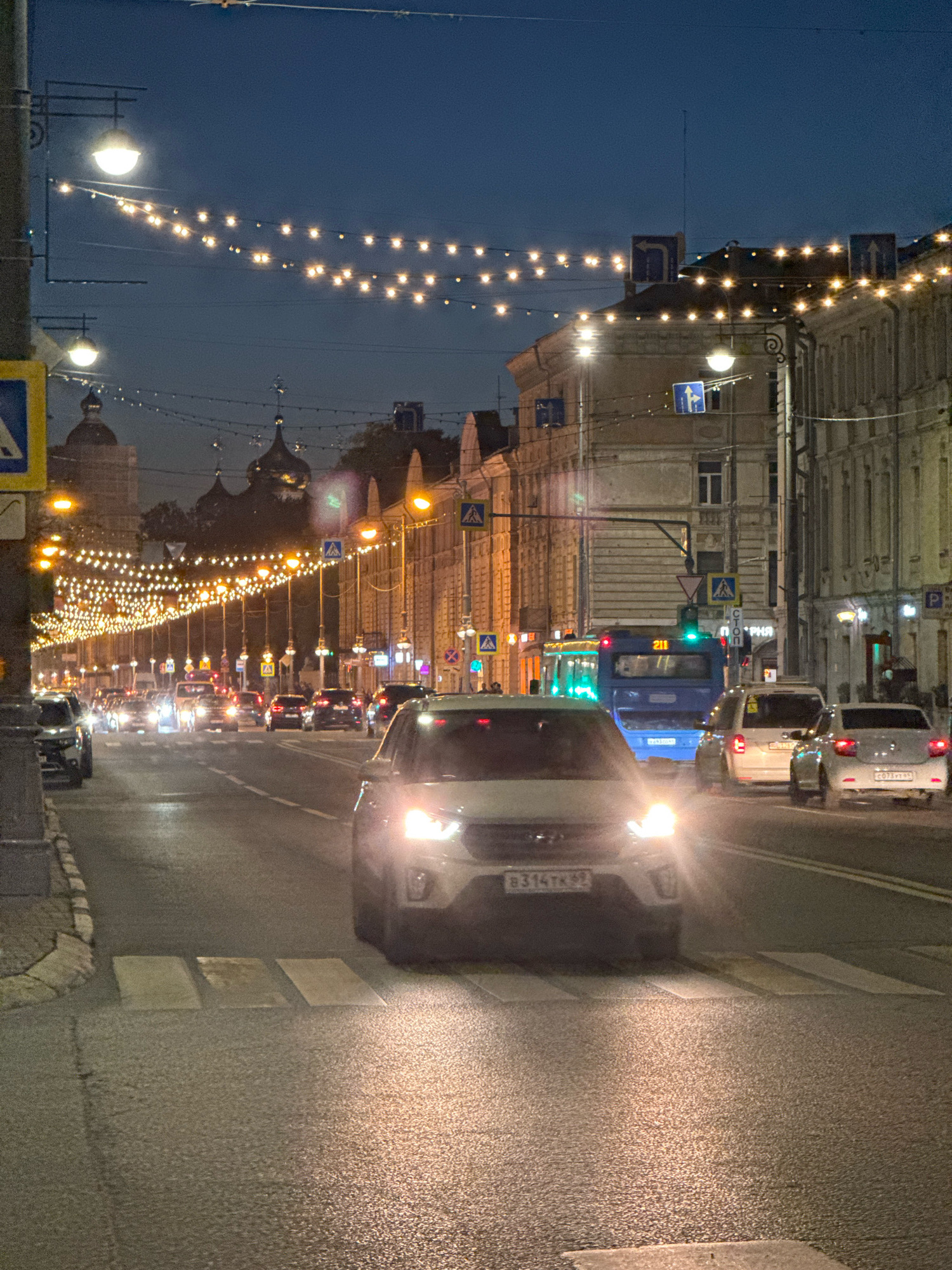 This is a night time street scene in Tver, Russia. The street is lined with buildings and illuminated by string lights and street lamps. Several cars and a bus are on the road, and a pedestrian crossing is visible in the foreground. The sky is dark blue, indicating it is either dusk or early evening. The overall atmosphere is festive due to the decorative lighting.