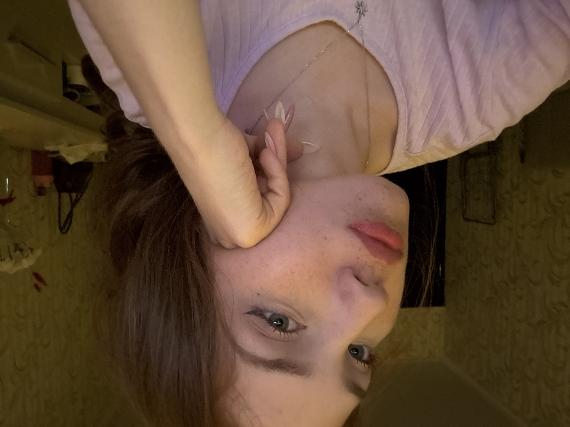 A young woman is depicted in an indoor setting, posing with her left hand supporting her chin and jawline. She is shown from the chest up, angled slightly to her right, with her head turned towards the viewer. Her skin is fair, with numerous freckles visible across her nose and cheeks. She has light-colored eyes, outlined with black eyeliner forming a wing, and her eyelashes appear enhanced with mascara. Her lips are full and coated with a pinkish-red lipstick. Her hair is dark brown and appears long, styled down, with some strands visible over her right shoulder. She is wearing a light pink, possibly ribbed or textured, short-sleeved top with a rounded neckline. A silver-colored necklace with a small, star-shaped pendant hangs around her neck. Her left hand displays long, manicured fingernails with a distinct white and pink ombre design, possibly artificial enhancements.
The background is an interior room with dim, warm, yellowish lighting. The wall behind her is covered with wallpaper featuring a repeating, subtly embossed, scroll or floral pattern in a muted yellow-brown hue. To the left, a dark vertical element, possibly a doorframe or furniture edge, is partially visible. To the right, a light-colored surface, resembling a desk or dresser, holds several objects, including a dark bag, an orange bottle, and some white materials. The overall scene suggests a residential indoor environment. The image contains no visual information that directly confirms the location as Naberezhnye Chelny, Russia.