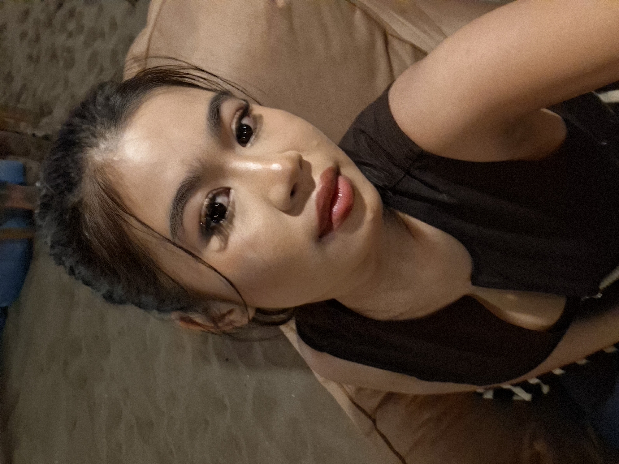 An Indonesian woman with dark hair pulled back in a bun and dark eye makeup is captured in a close-up, selfie-style shot. She is wearing a dark, sleeveless top with a V-neck opening and is leaning back on a large, light brown cushion. The background appears to be a sandy area, possibly a beach or outdoor seating area with sand on the ground. The lighting suggests it is evening or night, with warm, ambient light casting shadows. The setting is identified as Kuta, Indonesia.