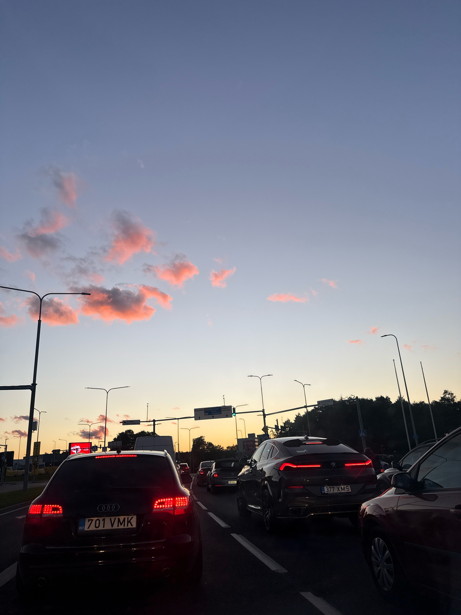 The image captures a traffic jam on a street in Tallinn, Estonia, during sunset. The sky is a gradient of soft blue transitioning to a pale yellow near the horizon, with wispy pink clouds scattered across it. Several cars are stopped in traffic, their brake lights illuminated. In the foreground, a black Audi station wagon with license plate "701 VMK" is visible. To its right, a dark-colored BMW SUV with license plate "377 XMS" is also stopped. Streetlights and traffic signals are present throughout the scene. Trees line the background, silhouetted against the twilight sky. A digital billboard with a red advertisement is visible in the distance on the left. The overall impression is a typical evening commute in an urban environment.