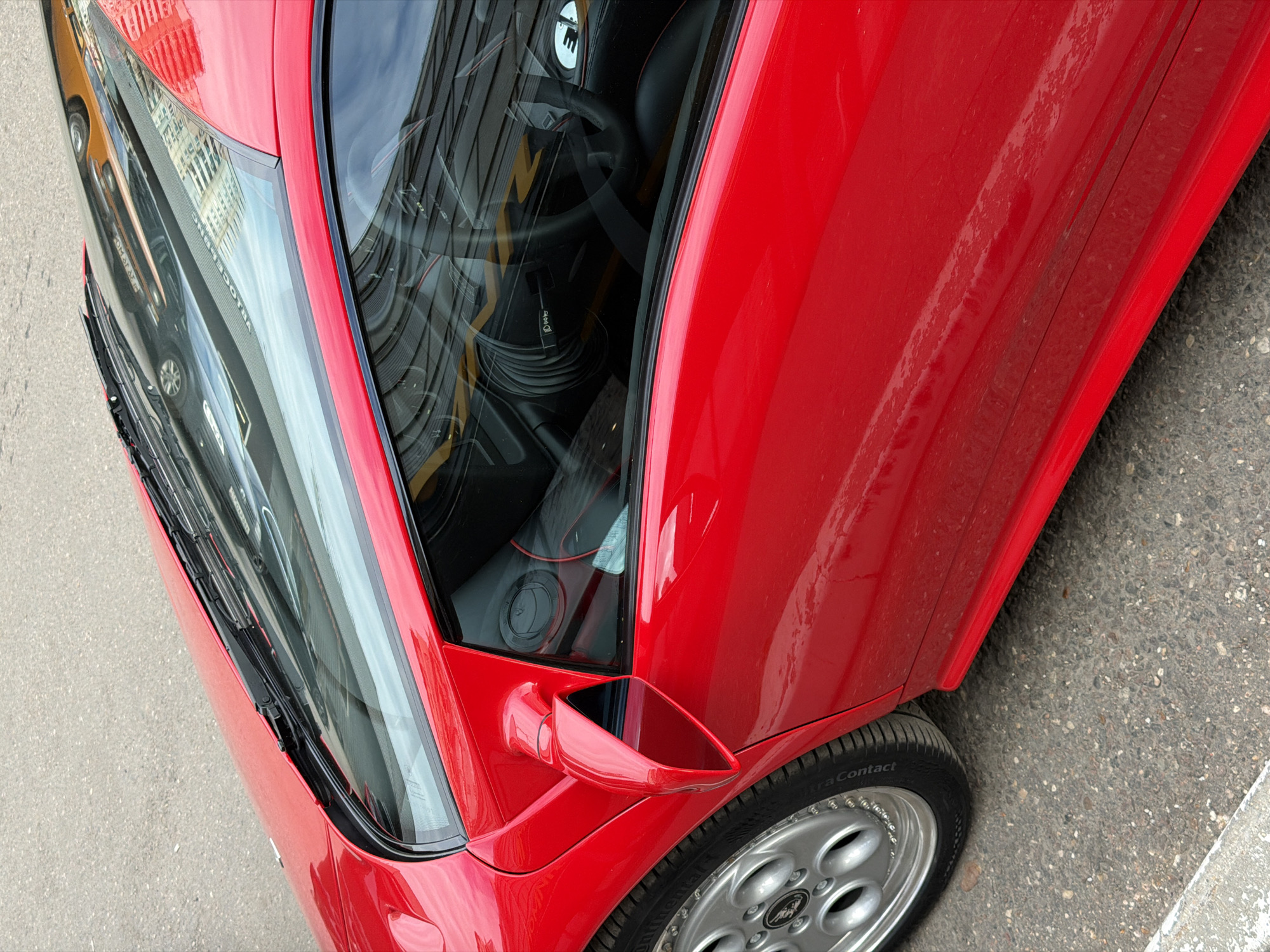The image, taken from an elevated perspective looking downwards, features a bright red Lamborghini Diablo sports car parked on an asphalt street in Moscow, Russia. The car is positioned at an angle, with its front and passenger side visible.
The polished red bodywork of the car reflects the surrounding environment, including a multistory building with numerous windows and a clear, blue sky. On the front windshield, mirrored and upside-down reflections clearly show the Cyrillic text "МОСКОВСКИЙ ПРОСПЕКТ" (MOSKOVSKY PROSPEKT), confirming the location as Moscow Avenue. The car's windshield wipers are visible at the base of the front glass.
Through the passenger side window, parts of the car's interior are visible. A black steering wheel can be seen, with a distinctive white circular emblem on its horn cap, which appears to be a stylized "I" with an inverted "V" shape, resembling an Iron Man logo.
The lower side panel of the red car shows some accumulation of dirt and dust, indicating it has been driven or parked outdoors. The red side mirror is angled outwards. A portion of the front right wheel is visible, featuring a silver, multi-spoke rim with the iconic Lamborghini bull logo at its center. The tire's sidewall displays the partial text "traContact," suggesting Continental UltraContact tires. The car is parked on coarse asphalt pavement, and a grey concrete curb is visible along the bottom right edge of the frame. The reflections suggest a clear and bright daytime setting.