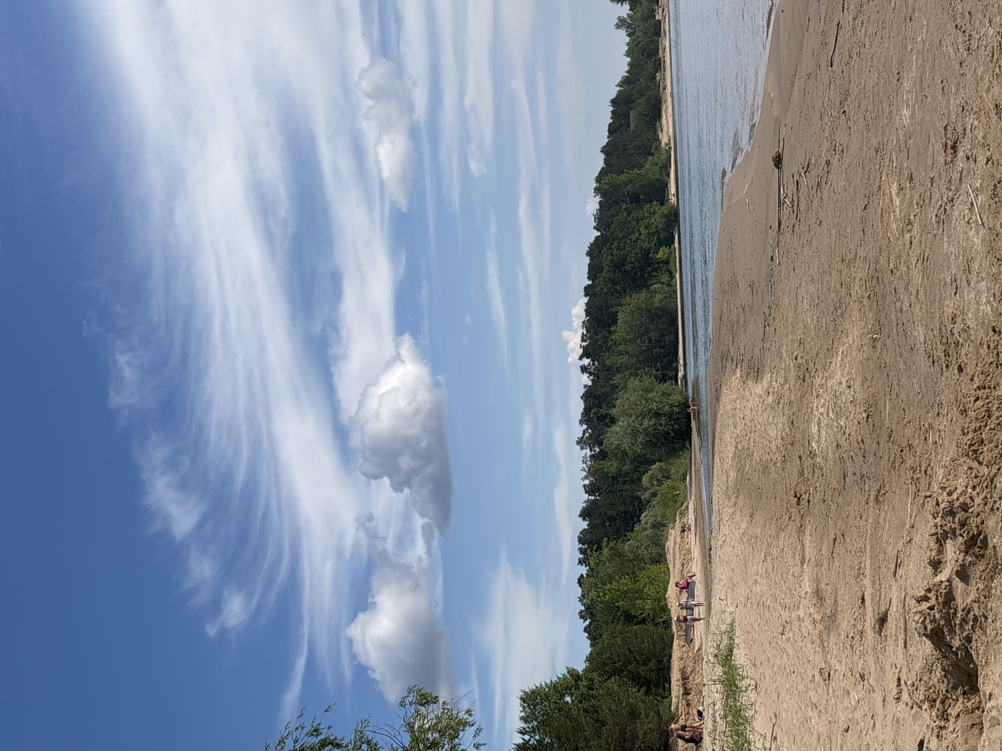 A wide shot of a sandy beach in Kzyl-Bairak, Russia, reveals a calm body of water on the right side and a dense forest in the background. Several people are scattered across the beach, some walking, some sitting, and others playing near the water's edge. The sky is a vibrant blue with streaks of white clouds. The sand is light brown and appears slightly damp in some areas closer to the water. Trees, predominantly coniferous, form a dark green line along the horizon, with some deciduous trees interspersed. The overall scene depicts a leisurely day at a natural outdoor setting.