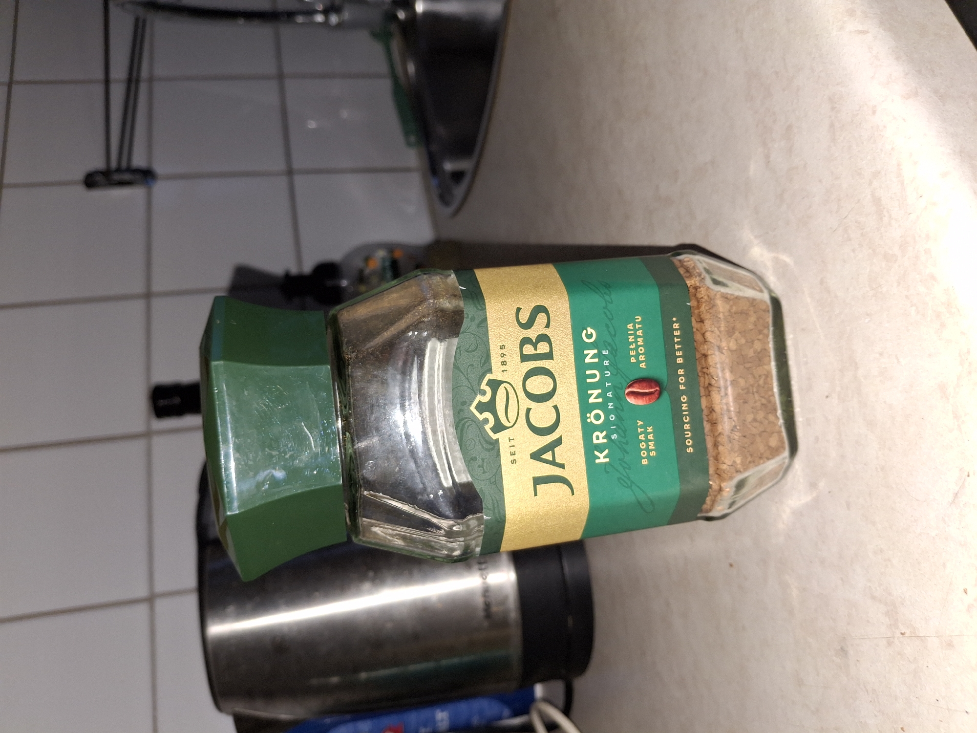 A jar of Jacobs Krönung instant coffee sits on a speckled countertop in a kitchen. The jar is mostly full of coffee granules. The label on the jar is gold and green with the Jacobs logo and "Krönung Signature" printed on it. To the left of the jar is a stainless steel kettle. In the background, there are white tiled walls and a faucet. The scene is illuminated by overhead lighting. The location of Åsgårdstrand, Norway is not discernible from the image.