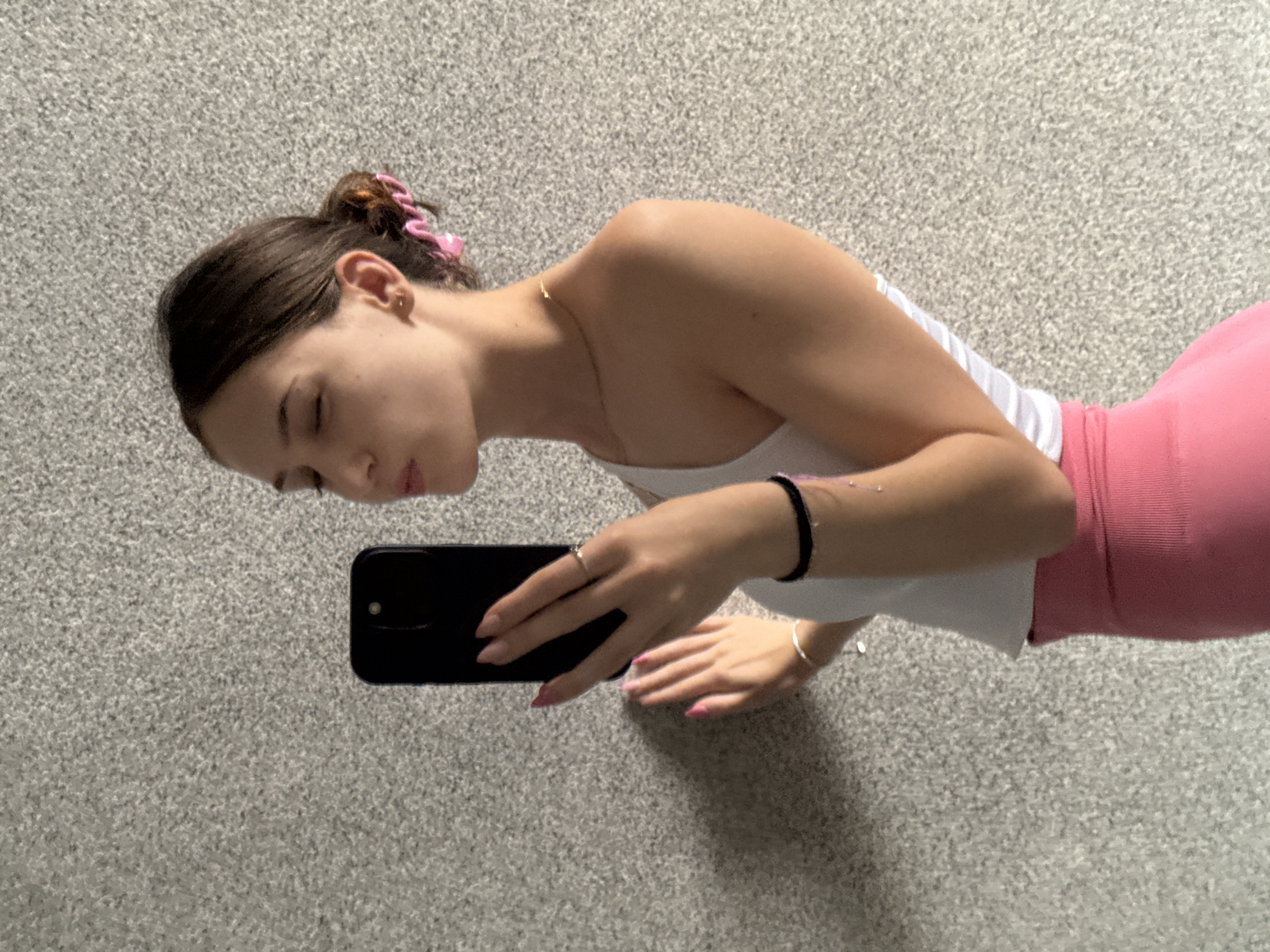 The image shows a young woman lying on a grey, carpeted floor, taking a selfie with her smartphone. She has dark hair pulled up in a bun with a pink scrunchie and is wearing a white tank top and pink leggings. Her right hand holds the phone, while her left arm rests on the floor. She is wearing a thin gold necklace, a black bracelet, and a silver ring. Her fingernails are painted pink. The lighting suggests it is daytime, likely indoors. The overall impression is casual and relaxed. The background is a textured grey carpet, providing no specific location cues beyond an indoor setting. There is no visible text in the image.