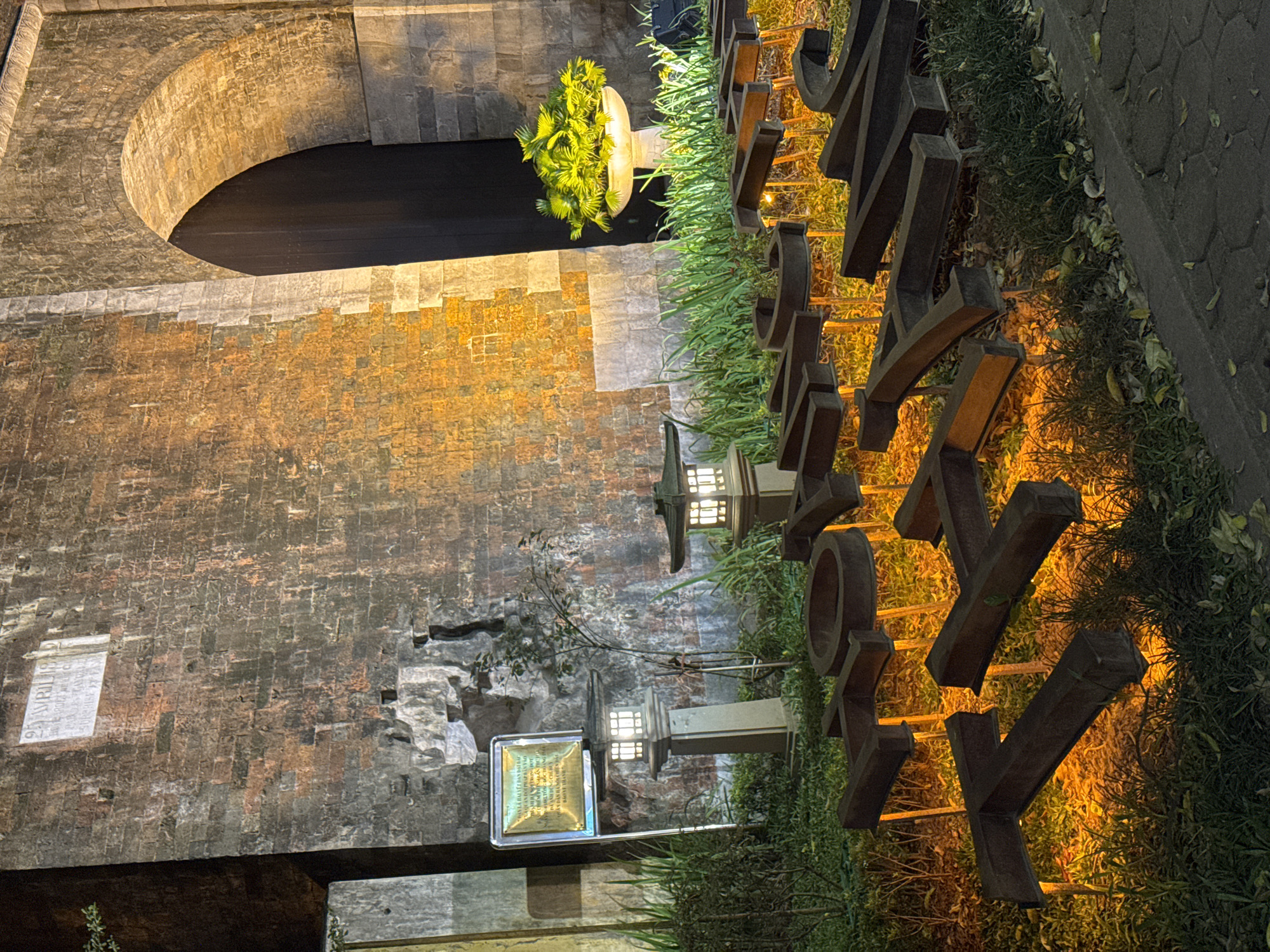 The image depicts a nighttime scene in Hanoi, Vietnam, featuring a large brick archway with a dark opening. The brickwork of the arch is illuminated, casting a warm, orange glow. To the right of the arch, a large, three-dimensional metal sign with dark, block letters spelling "HANOI" is prominently displayed. The letters are illuminated from behind, creating a soft, ambient light that highlights the surrounding greenery.
The sign is surrounded by lush green plants and grass, which are also lit by warm, yellow lights, giving the scene a vibrant and inviting atmosphere. Two traditional-style lanterns with white frosted glass panels are placed along the path in front of the sign. The ground to the right of the sign appears to be paved with hexagonal stones.
On the brick wall to the left of the arch, there is a white rectangular plaque with visible text. Another plaque, this one metallic and rectangular, is mounted on a pole near the lanterns, also containing text. The overall impression is one of a well-lit, possibly public space in Hanoi at night, emphasizing both historical architecture and modern signage. The lighting suggests a curated and aesthetically pleasing environment.