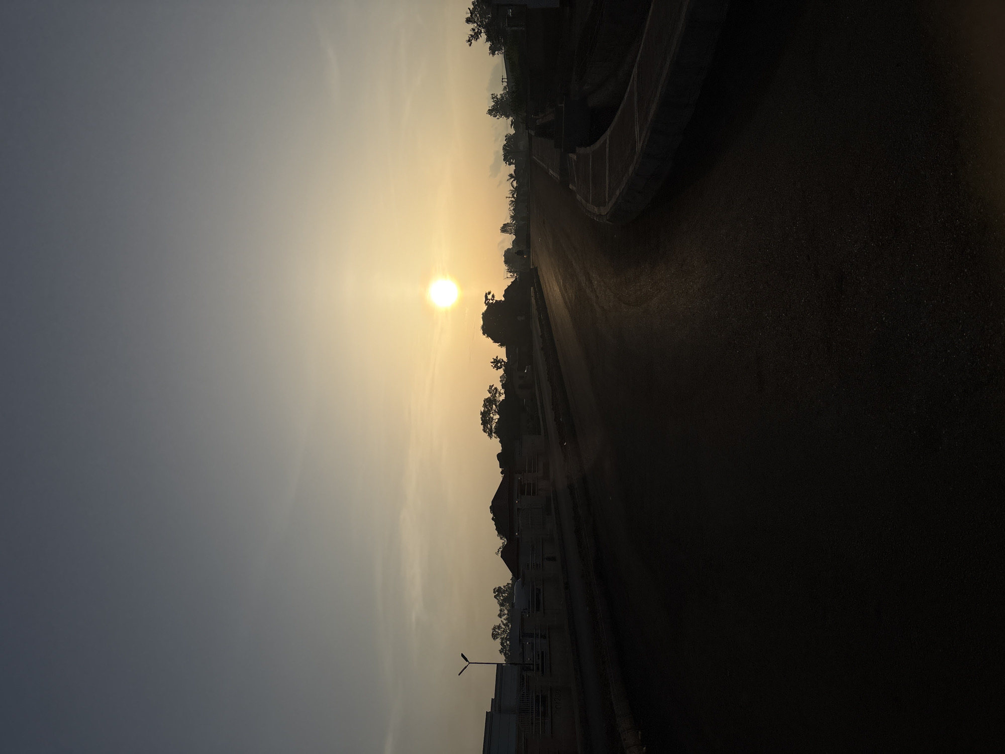 The image shows a sunrise over a road in Bukuru, Nigeria. The sun is a bright, yellow orb low in the sky, casting a warm glow. The sky is a gradient of pale yellow and blue. A paved road curves into the distance, with a concrete barrier on the right side. Silhouettes of trees and buildings are visible along the horizon. The foreground of the road is dark and indistinct. A lamppost stands to the left of the road, and a fence with metal railings borders the properties on the left. The overall scene depicts an early morning in a developing area, with the natural beauty of the sunrise contrasting with the urban landscape.