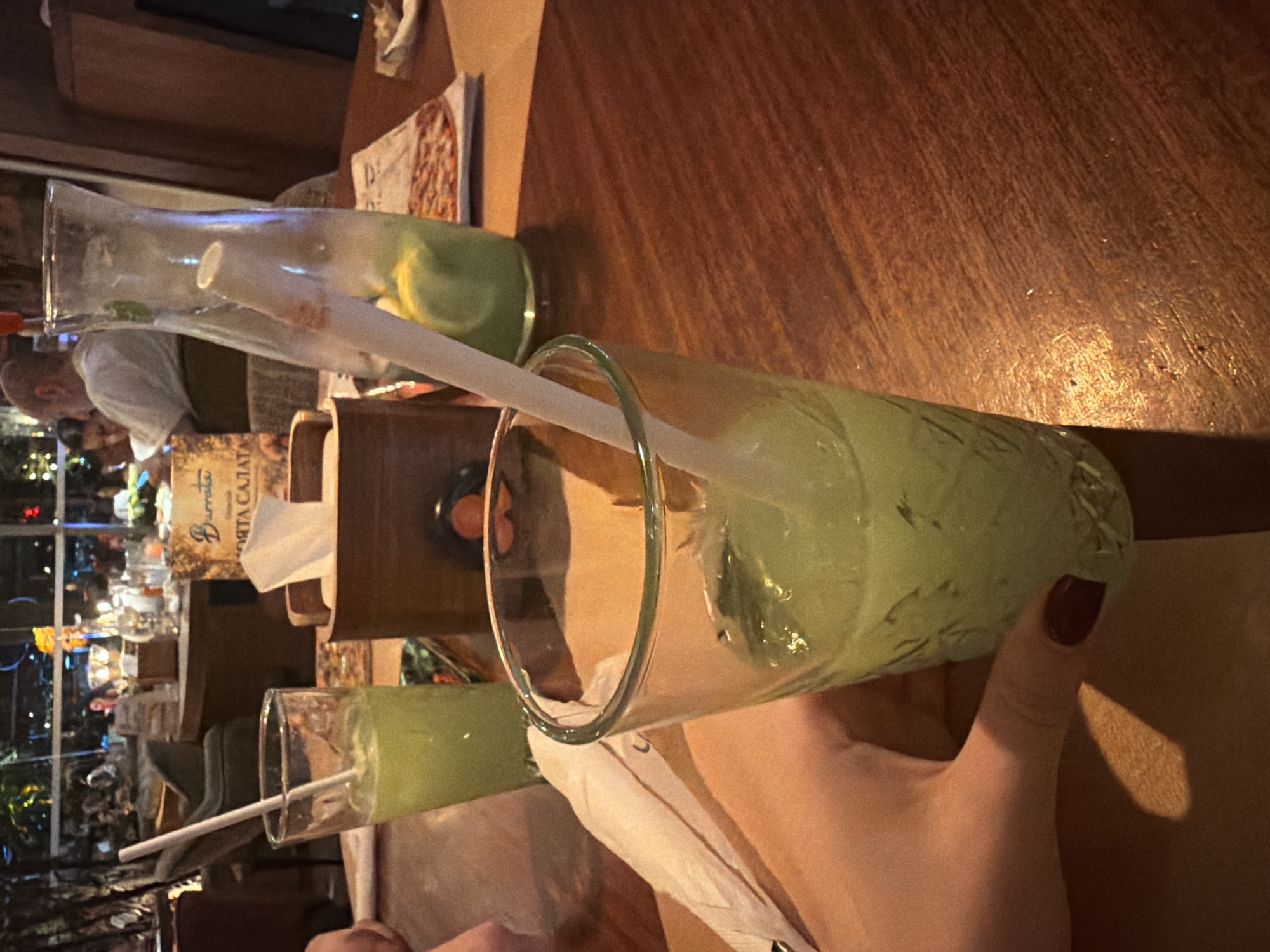 An indoor restaurant scene is depicted at night, characterized by warm, dim lighting. In the foreground, a light-skinned hand with dark red nail polish on the thumb holds a textured clear glass containing a light green beverage and a white straw. To the immediate left, another similar glass, partially obscured, also holds the green beverage and a straw. A folded white paper napkin rests beside it.
The drinks are placed on a dark wooden table with visible grain. In the midground, a clear glass pitcher, nearly empty of the same green beverage, contains ice, lemon slices, and a white straw. An open menu, featuring an image of pizza, lies on the table next to the pitcher. A wooden caddy, possibly for napkins, is also present. Behind it, a brown sign prominently displays "Burrata" in a script font, with Cyrillic text "МЕНЮ САЛАТА" (MENU SALADS) beneath, indicating a location where Bulgarian is spoken, such as Sofia, Bulgaria.
The background reveals a bustling restaurant environment with several patrons seated at tables. A bald man in a white shirt is clearly visible in the mid-background. Beyond the interior, large windows offer a blurred view of city lights at night. The overall ambiance is relaxed and social.