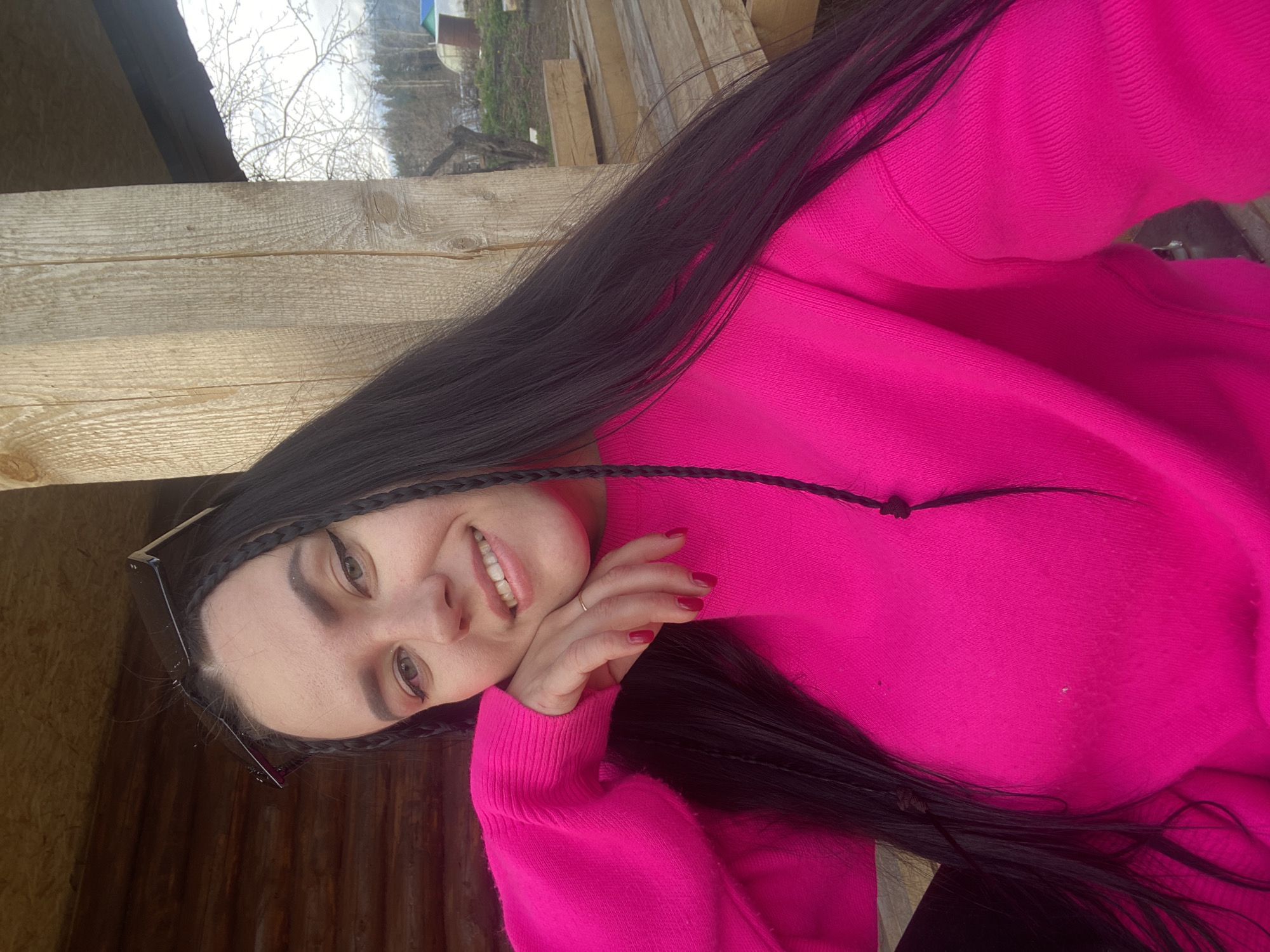 A woman with long, dark hair, styled with braids, is posing for a selfie. She is wearing a bright pink sweater. She is smiling. The background appears to be a wooden structure, possibly a shed or a building under construction, with some greenery visible in the distance. The location is Chaikovsky, Russia.