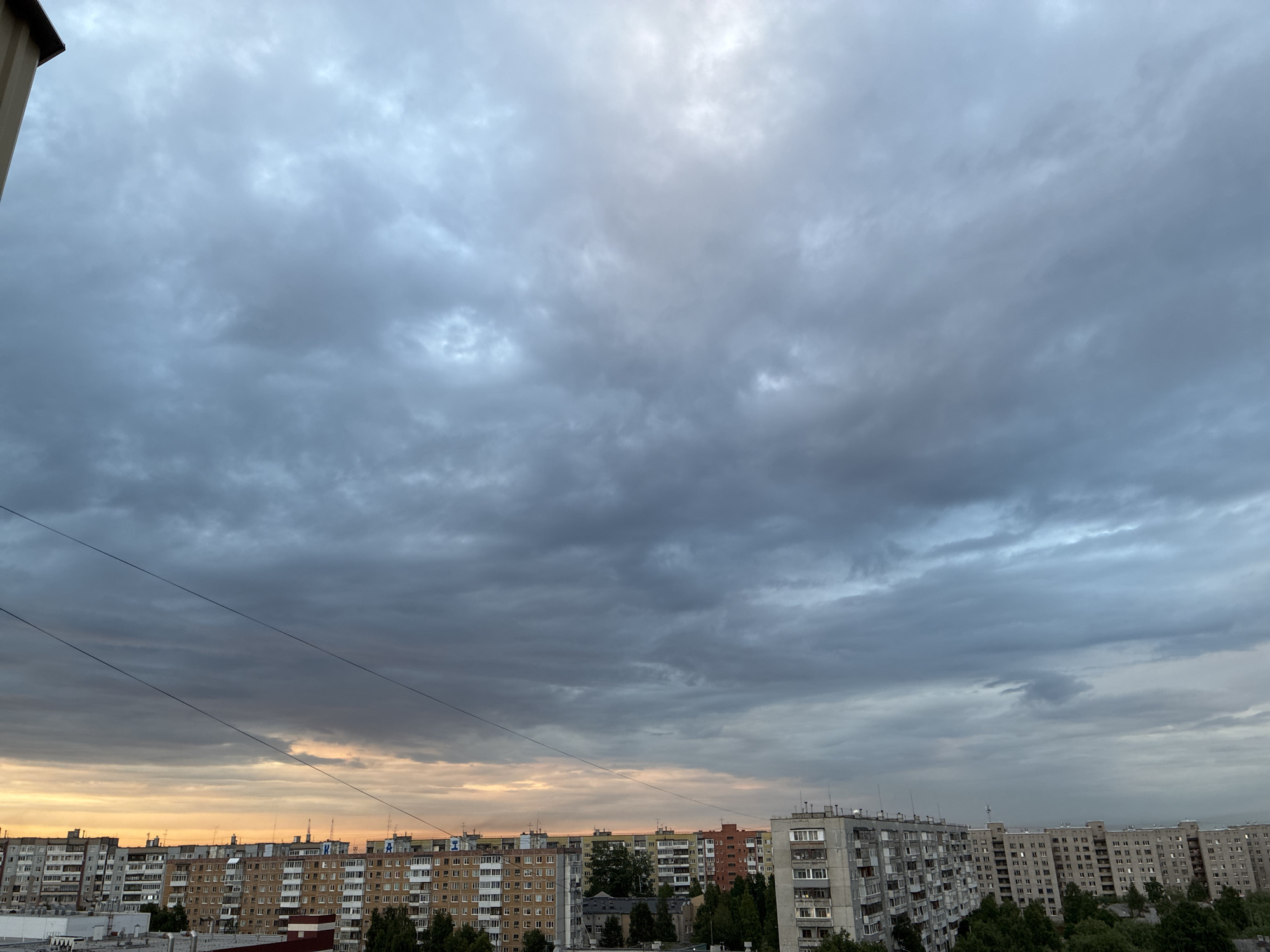 The image shows a cityscape at dusk. The sky is filled with heavy, grey clouds, with a faint orange glow on the horizon, indicating the setting sun. In the foreground, there are several multi-story apartment buildings, typical of Soviet-era architecture, lined up across the frame. Some of the buildings are lighter in color, while others are a darker brick. Power lines are visible stretching across the sky. The overall impression is a somewhat overcast and perhaps slightly gloomy urban scene.