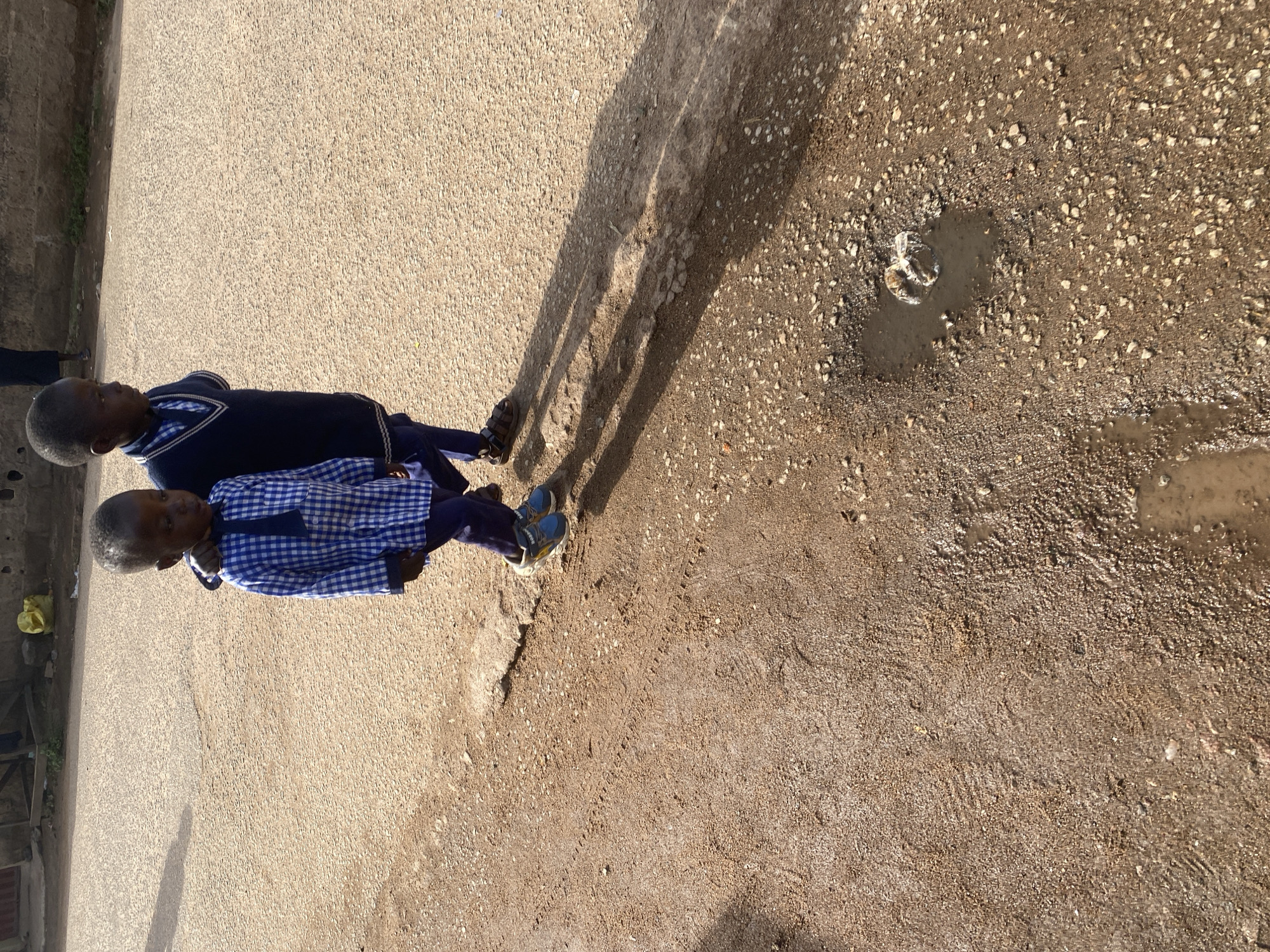 In the city of Jos, Nigeria, two young boys are walking on a paved road. The boy in front is wearing a blue and white checkered shirt and dark blue pants, with blue and grey sneakers. The boy behind him is wearing a dark blue v-neck sweater over a collared shirt, and dark blue pants, with brown sandals. They are casting long shadows on the road, indicating it is likely mid-day. The road is made of coarse, light brown gravel and asphalt. There are several small puddles of water on the road, one of which has a small, white object floating in it. In the background, a yellow plastic bag is visible on the side of the road, near a low wall made of rough, grey stones. A building structure is also visible in the background.