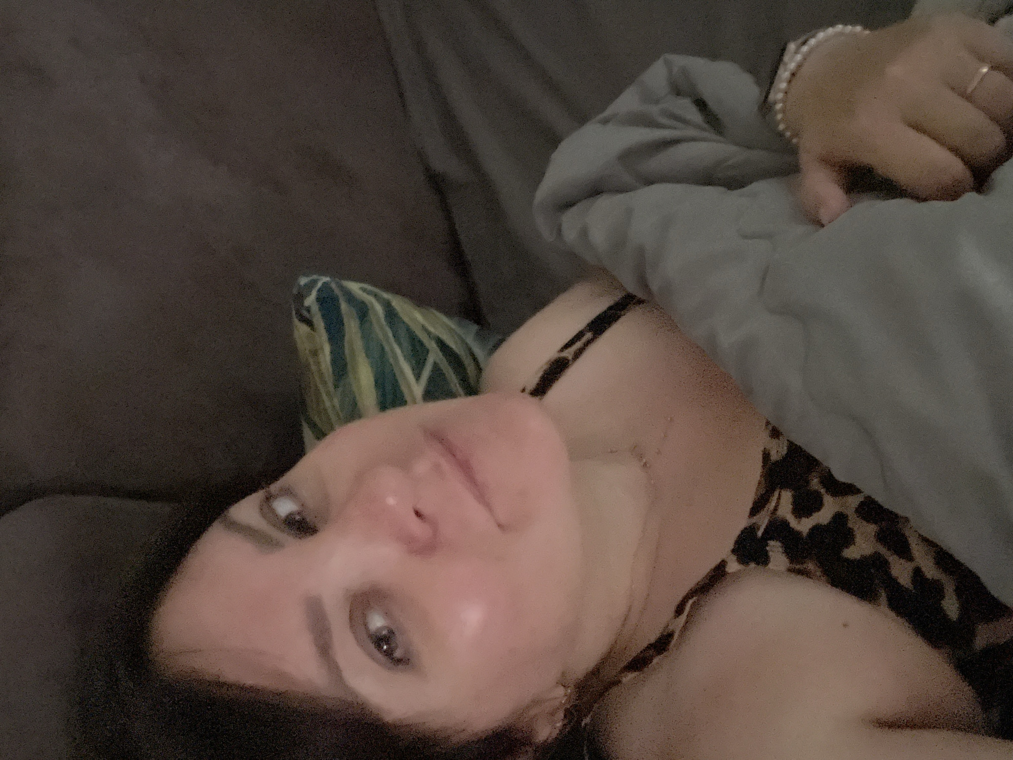 A woman with brown hair and brown eyes is lying down, holding a gray blanket. She is wearing a leopard-print top and a necklace. There is a pillow with a blue and green striped pattern behind her. The background appears to be a couch or bed. The image is a selfie, taken from a high angle.