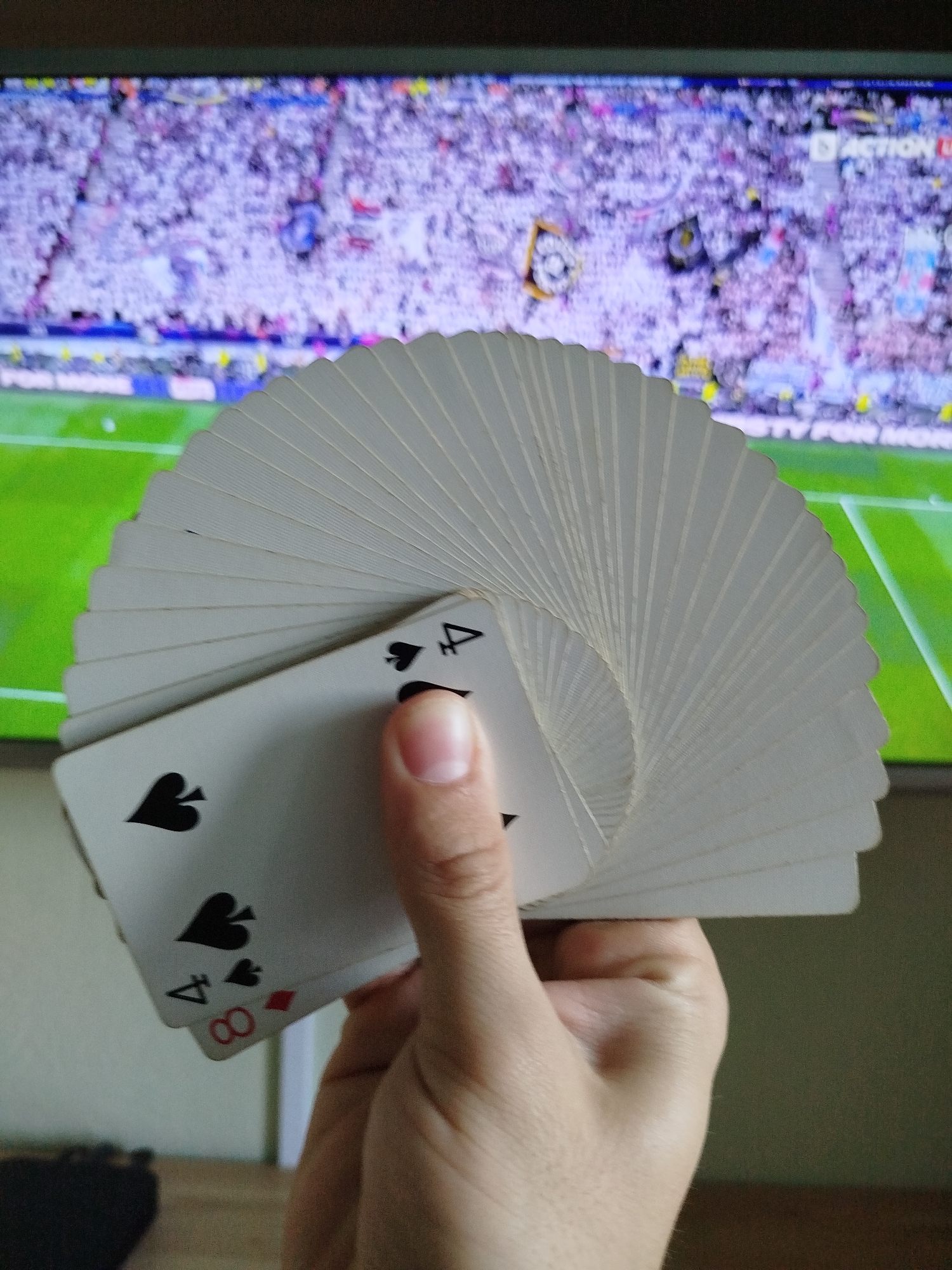 A hand holds a fanned deck of playing cards. The top card showing is the Ace of Spades, followed by the 4 of Spades, and partially visible beneath is the 8 of Diamonds. In the background, a television displays a soccer match with a large crowd. The location is not specified but does not appear to be in the city of Varna, Bulgaria.