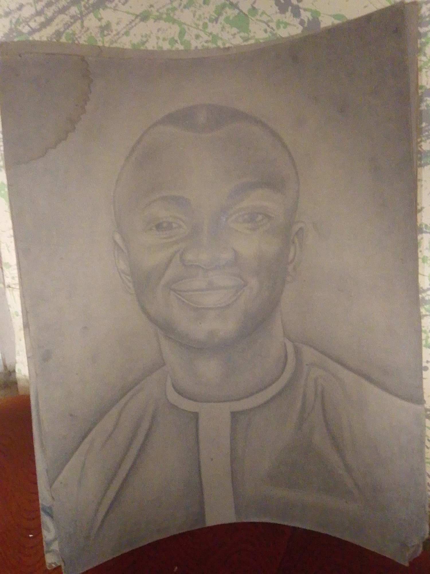This is a photograph of a charcoal sketch of a young man's head and upper torso. The sketch is on a light-colored paper with some staining and wear, particularly a large stain on the upper left corner. The man has a shaved head with a subtle part down the middle, prominent ears, and is smiling with his mouth slightly open. He is wearing a shirt with a rounded neckline and what appears to be a central placket. The background of the sketch is a gradient of gray tones, and the paper is placed on a surface that appears to be a wooden table or furniture. The overall lighting is subdued. The image is taken from a slightly elevated angle, and a portion of what looks like a patterned wallpaper or fabric is visible at the top of the photograph. There is no identifiable information suggesting the location is Jalingo, Nigeria.