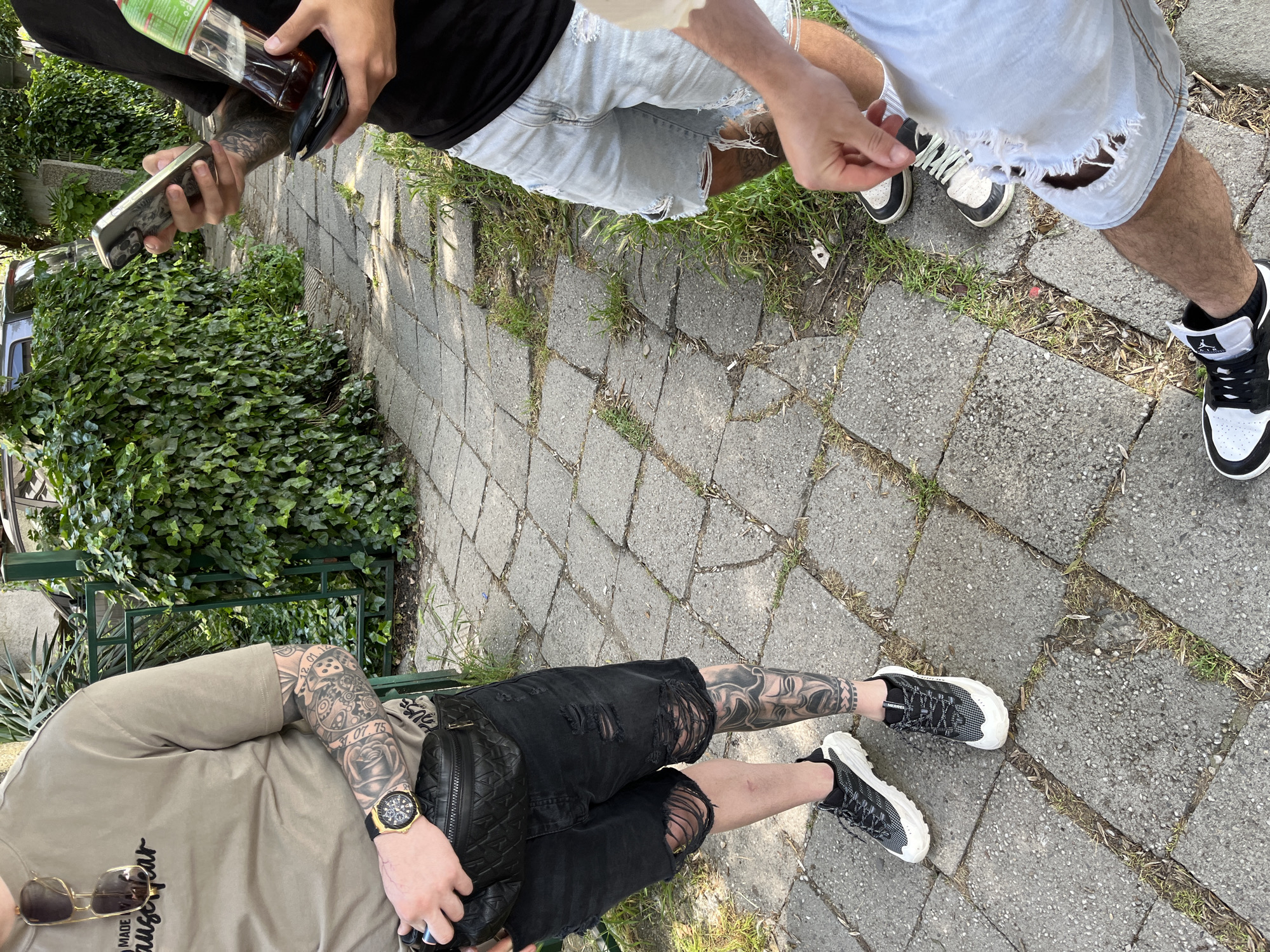 An overhead, slightly angled outdoor shot captures three individuals casually standing on a paved pathway. The scene is illuminated by bright daylight, with shadows indicating the sun is overhead. The ground is composed of rectangular concrete pavers, with grass and weeds growing in the gaps. To the left, a dense mass of green ivy covers a fence or wall, behind a green metal railing.
In the upper left, one person, wearing a black t-shirt, holds a silver-cased smartphone in their left hand and a clear glass bottle, partially revealing a green "Sprite" label, along with a black wallet in their right hand. Their forearm is adorned with intricate tattoos.
To the upper right, another person wears light blue distressed denim shorts with large rips and black and white high-top sneakers, featuring the "Air Jordan" logo. A hand is visible, adjusting a shoelace.
In the lower part of the frame, a third person is seen from the torso down. They are wearing a tan t-shirt with partial text "MADE IN HANGER," black distressed denim shorts, and black and white patterned sneakers. Extensive tattoos cover their left arm and legs, including designs of a rose, gears, and dates like "31.07.75" and "01.01.". A prominent gold-colored wristwatch is on their left wrist, and sunglasses hang from their t-shirt collar. They hold a black, quilted fanny pack.
The individuals appear to be in a relaxed, casual gathering or pause. A partial view of a silver vehicle can be seen through the foliage in the background. The setting suggests an urban or suburban path, consistent with the location context of Burgas, Bulgaria.
