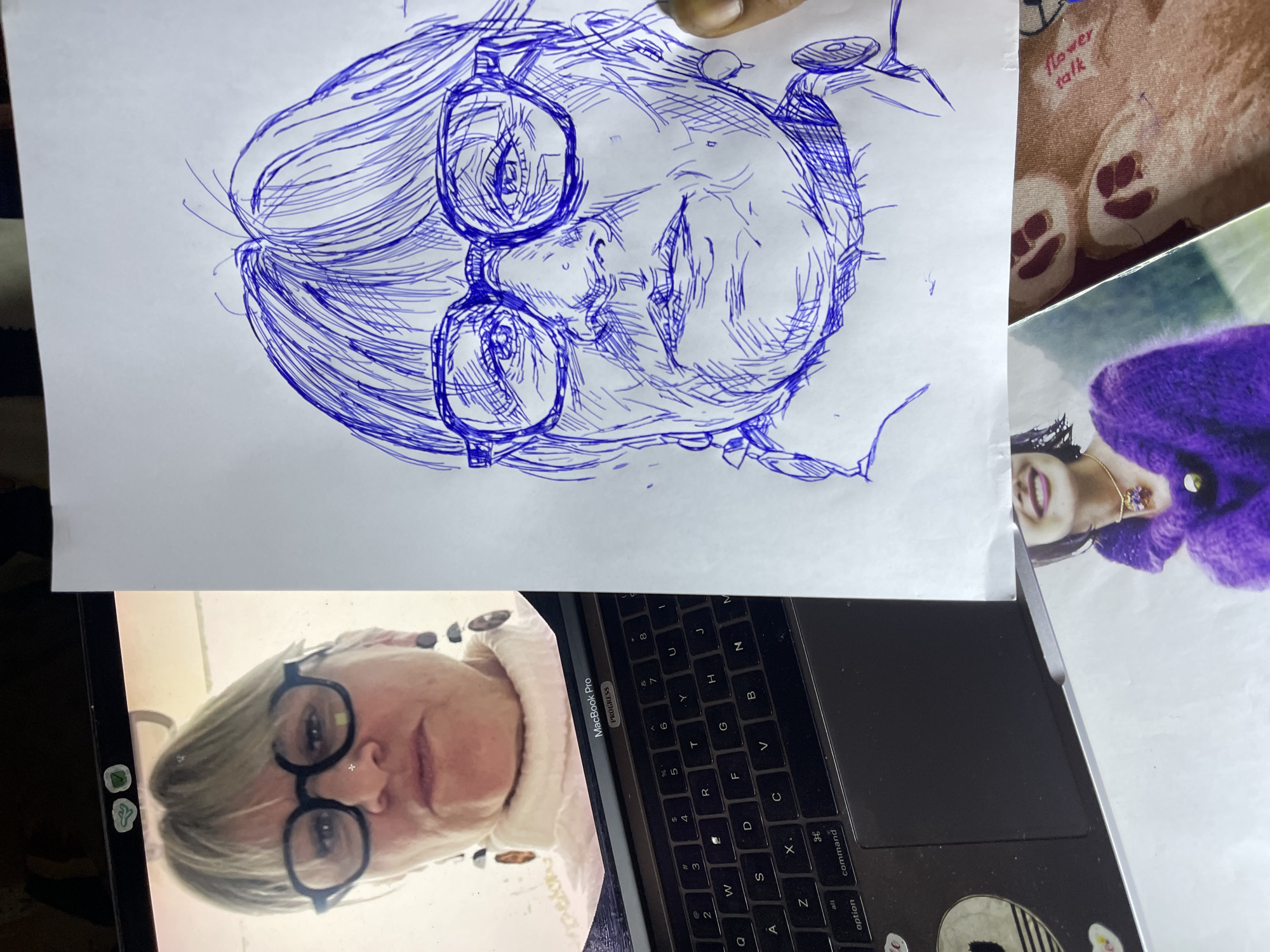 The image displays a juxtaposition of a digital portrait of a woman on a laptop screen and a hand-drawn sketch of the same woman. The laptop, a MacBook Pro, is partially visible in the foreground, its keyboard and trackpad in clear view. The screen shows a headshot of a woman with blonde hair, wearing large, dark-rimmed glasses and a white turtleneck. She has dangly earrings. To the right of the laptop, a sheet of white paper features a blue ink sketch of the woman, capturing her features with a focus on her glasses, eyes, and facial lines. The sketch is in progress, with some areas more detailed than others. Below the sketch, a photograph of a smiling woman wearing a purple fuzzy sweater and a necklace is partially visible. The scene is set against a backdrop of various textures, including what appears to be fabric with a teddy bear pattern and the word "flower talk" printed on it. The overall impression is of an artistic process, with a reference image being translated into a sketch. There is no indication of the city of Jos, Nigeria, or any specific urban environment.