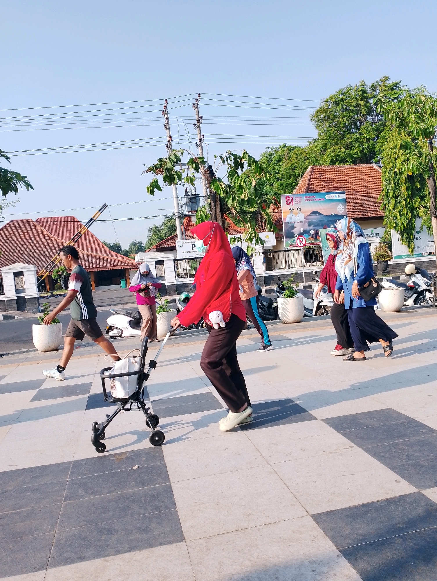 The image depicts a pedestrian walkway in Probolinggo, Indonesia, on a sunny day with a clear blue sky. Several individuals are walking along the tiled path, which features a checkerboard pattern of light and dark grey tiles.
In the foreground, a person in a red hijab and mask is walking with a stroller. To their left, a man in a dark t-shirt with white and red accents and dark shorts is walking, also in motion. Further behind them, two women, both wearing hijabs, are walking together. The woman on the left wears a maroon top and beige pants, while the woman on the right wears a blue top and a dark skirt, carrying a black bag. Another person, partially visible in the left background, is wearing a light purple hijab and pink shirt.
The environment includes potted plants, a white scooter, and motorcycles parked along the side of the walkway. In the background, there are buildings with red tiled roofs, a banner with Indonesian text and imagery, and utility poles with electrical wires. The overall scene suggests a public space being utilized for recreation or daily transit.