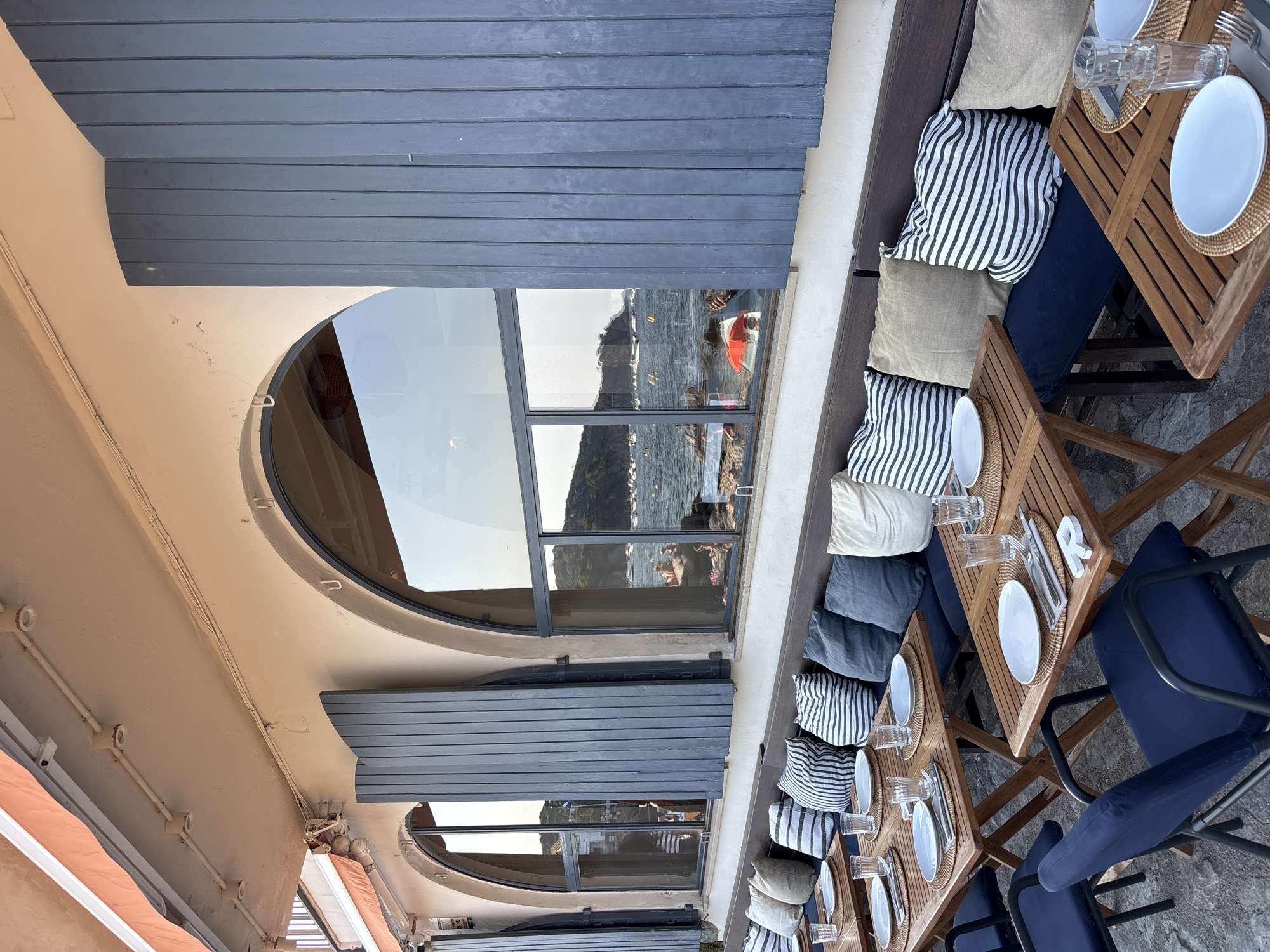 The image depicts an outdoor seating area of a restaurant in Calella De Palafrugell, Spain, overlooking the Mediterranean Sea. Several wooden tables are set with white plates, cutlery, and glasses on woven placemats. Dark blue chairs with metal frames surround some of the tables. A long, built-in wooden bench runs along the wall, adorned with a collection of striped and plain cushions in shades of blue, white, and beige. The seating area is covered by a pale orange awning, and large, arched windows with dark grey wooden shutters are visible. Through the windows, the sea can be seen with boats and a distant coastline. The overall scene conveys a relaxed, seaside dining atmosphere.