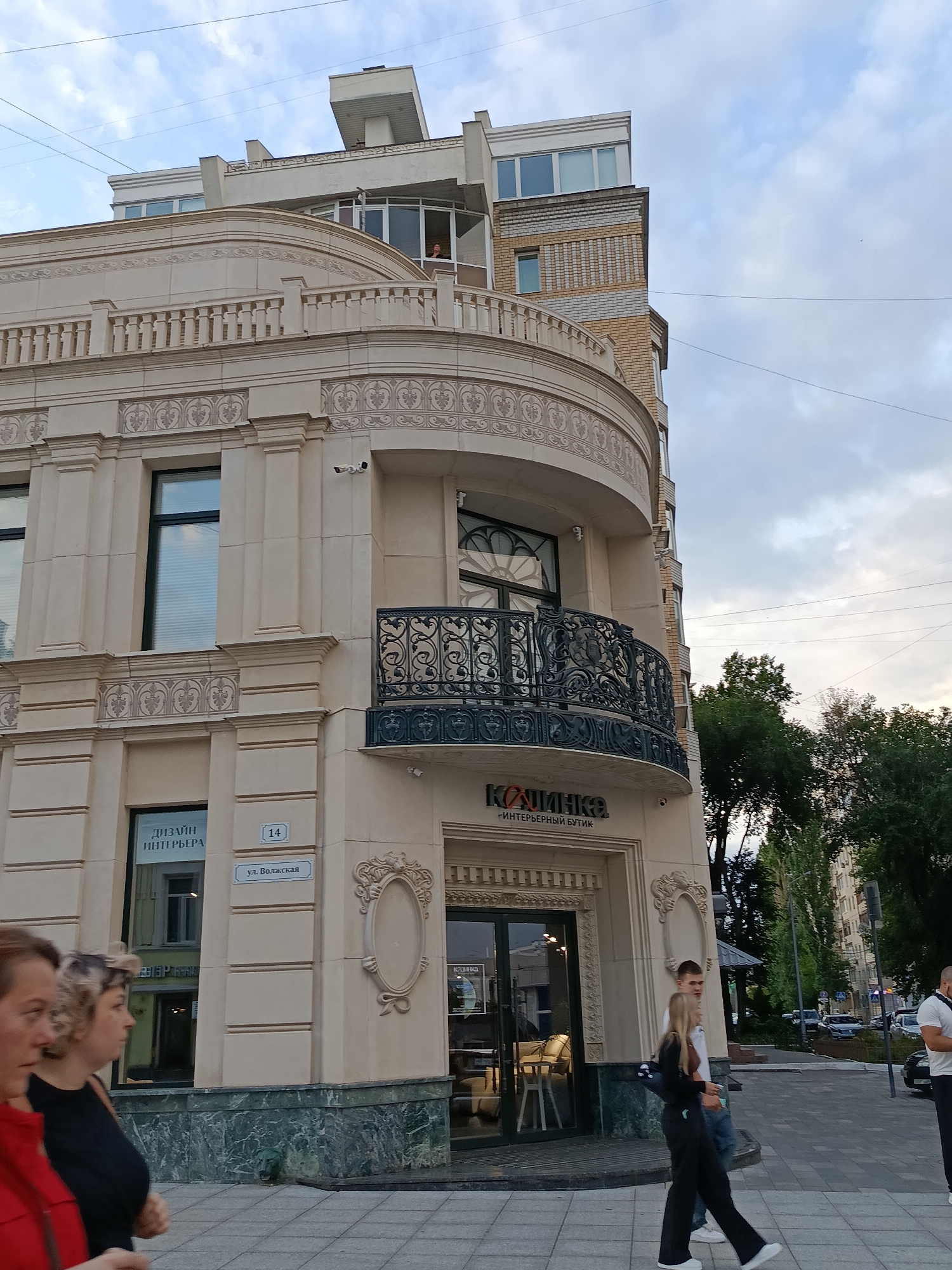 The image captures a street view of a corner building in Saratov, Russia, featuring a blend of modern and classical architectural elements. The building's facade is adorned with decorative moldings and columns, with a curved balcony on the second story featuring an ornate wrought-iron railing. A sign above the entrance reads "КАЛИНКА" and below it "ИНТЕРЬЕРНЫЙ БУТИК," indicating an interior design boutique. Above this, a sign for "ДИЗАЙН ИНТЕРЬЕРА" is visible. A street sign indicates "ул. Волжская" (Volzhskaya Street) and the number "14."
In the foreground, two women are walking from left to right. The woman on the left is wearing a red jacket, and the woman on the right has blonde hair and is wearing a black top and pants. Further to the right, a couple, a man and a woman, are also walking. The man is wearing a white shirt and jeans, and the woman is wearing a white top and black pants. In the background, trees and other buildings line the street, and cars are visible on the road. The sky is partly cloudy. The overall scene depicts an urban environment during daylight hours.