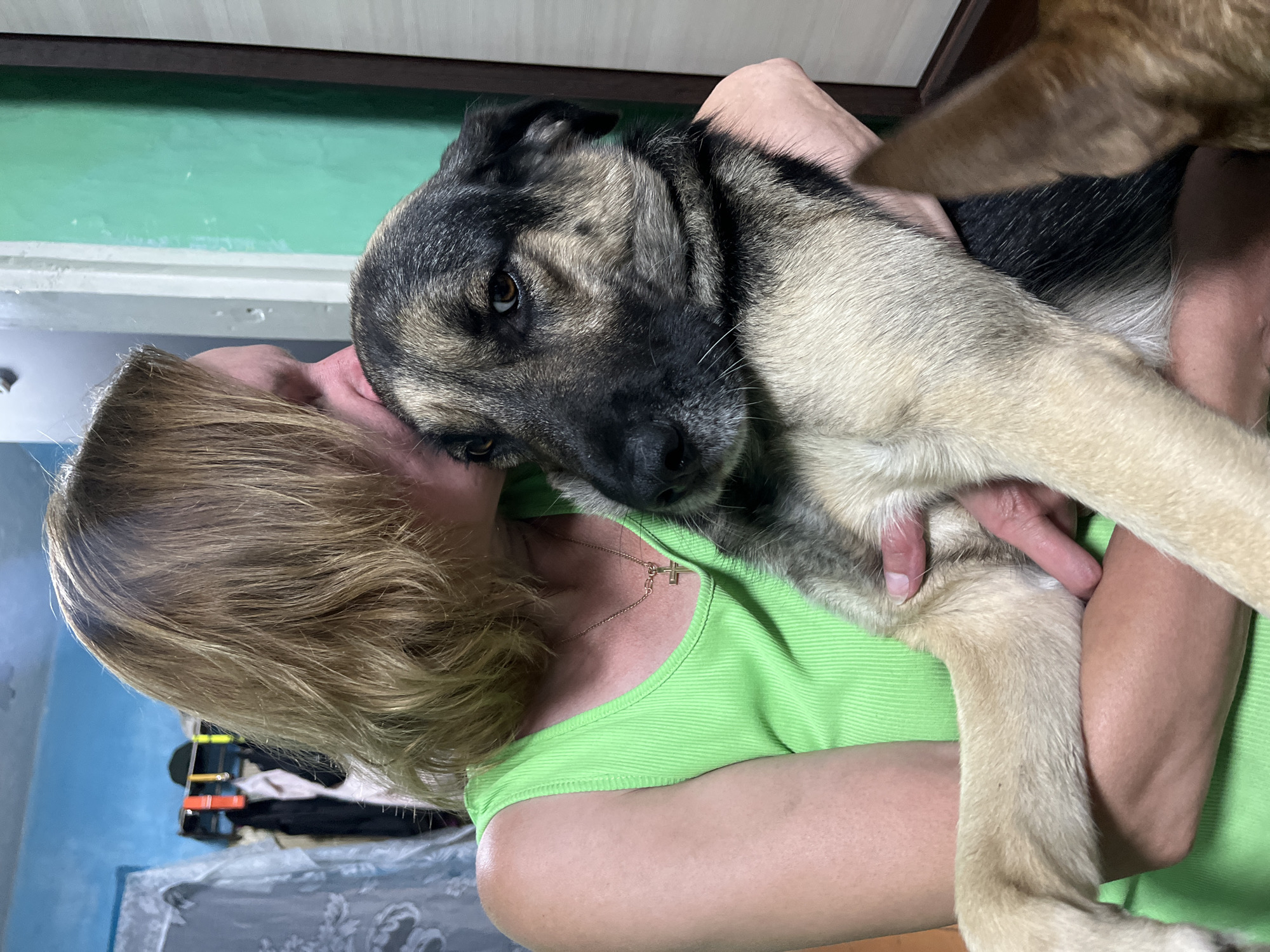 A woman with blonde hair, wearing a lime green tank top and a gold cross necklace, is holding a medium-sized dog with tan and black fur. The woman is kissing the top of the dog's head. The dog's face is visible, and it appears to be looking slightly to the right. Another dog's ear, brown in color, is visible in the lower right corner of the frame. The background is blurry and appears to be indoors, with a light blue wall with clothing on hangers to the left and a green wall to the right. The lighting suggests an indoor environment. The scene is located in Nizhny Tagil, Russia.