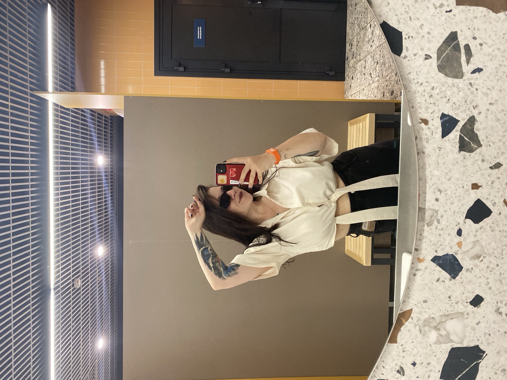 A woman with dark hair, wearing sunglasses, a white cropped shirt tied at the waist, and black pants, is taking a mirror selfie in what appears to be a changing room or public restroom. She has tattoos on her left arm and a crescent moon tattoo on her right wrist. She is holding her phone in her right hand and has her left arm raised above her head. The background shows a beige wall, a yellow tiled section, a dark door with a blue sign, and a bench. The foreground features a patterned mirror. The image is taken in a public facility.