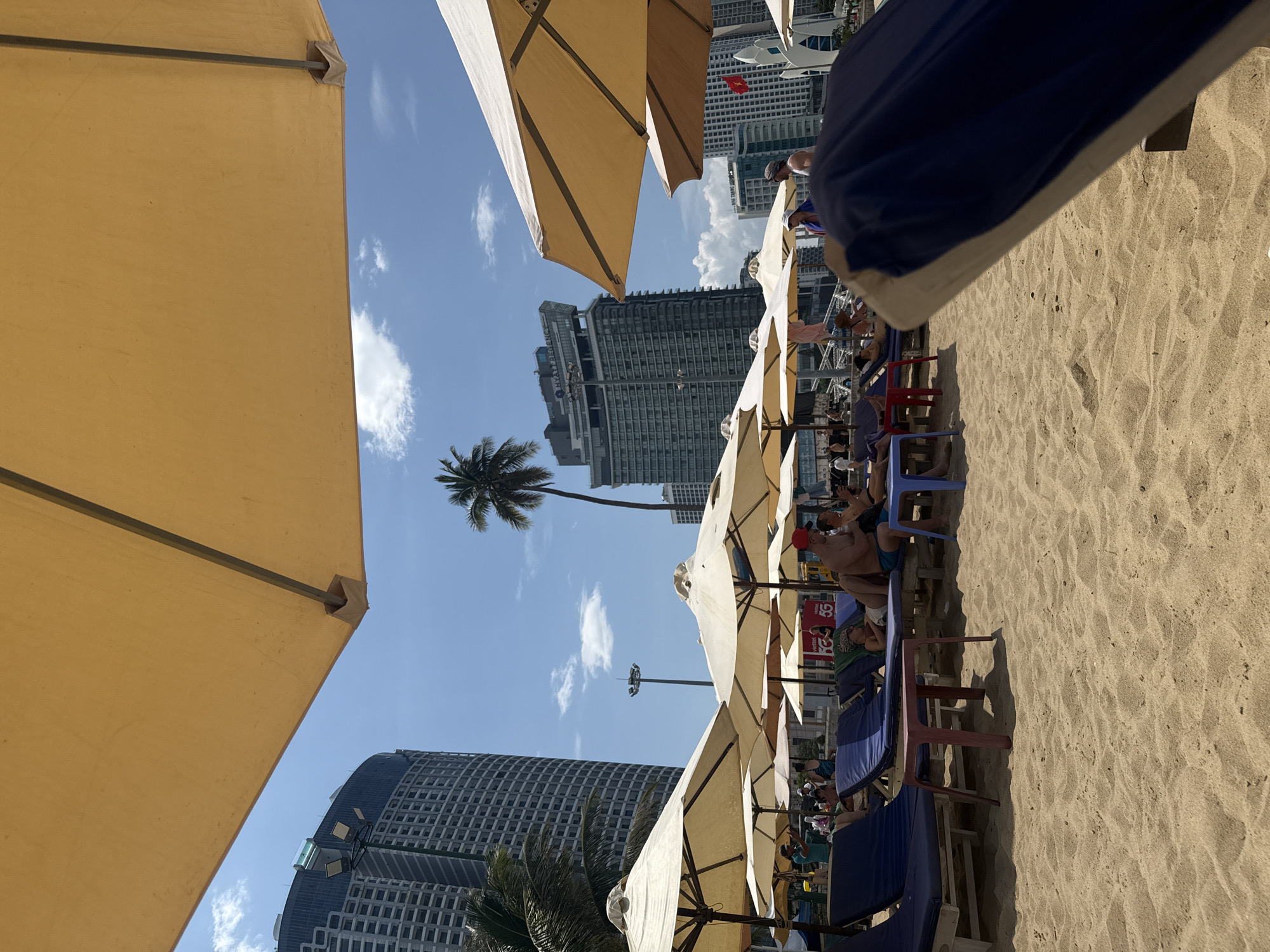 The image shows a beach scene in Nha Trang, Vietnam, with several rows of empty and occupied lounge chairs under large yellow umbrellas. In the background, tall buildings and palm trees are visible under a clear blue sky with scattered clouds. People are scattered among the lounge chairs, some are lying down, while others are sitting up and conversing. A few small tables are placed between the chairs. The sand is light brown and appears well-maintained. The overall impression is a sunny day at a popular tourist destination, likely on Nha Trang's main beach strip.