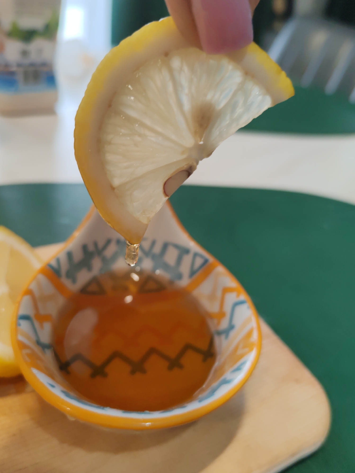 A slice of lemon is held by a person's fingers over a small, decorative bowl filled with amber-colored liquid, likely honey. A single drop of liquid is suspended mid-air as it falls from the lemon into the bowl. The bowl has an orange rim and a patterned exterior featuring zigzags and triangles in orange, white, and teal. A second, partially visible slice of lemon rests to the left of the bowl. The subjects are positioned on a light-colored wooden surface, with a green surface visible in the background. The lighting appears bright, casting subtle reflections on the liquid and the lemon. The scene is captured in a close-up, shallow depth-of-field, focusing attention on the dripping lemon and the bowl of honey. There is no discernible information about the location in Komsomolsk-on-Amur, Russia, within this image.