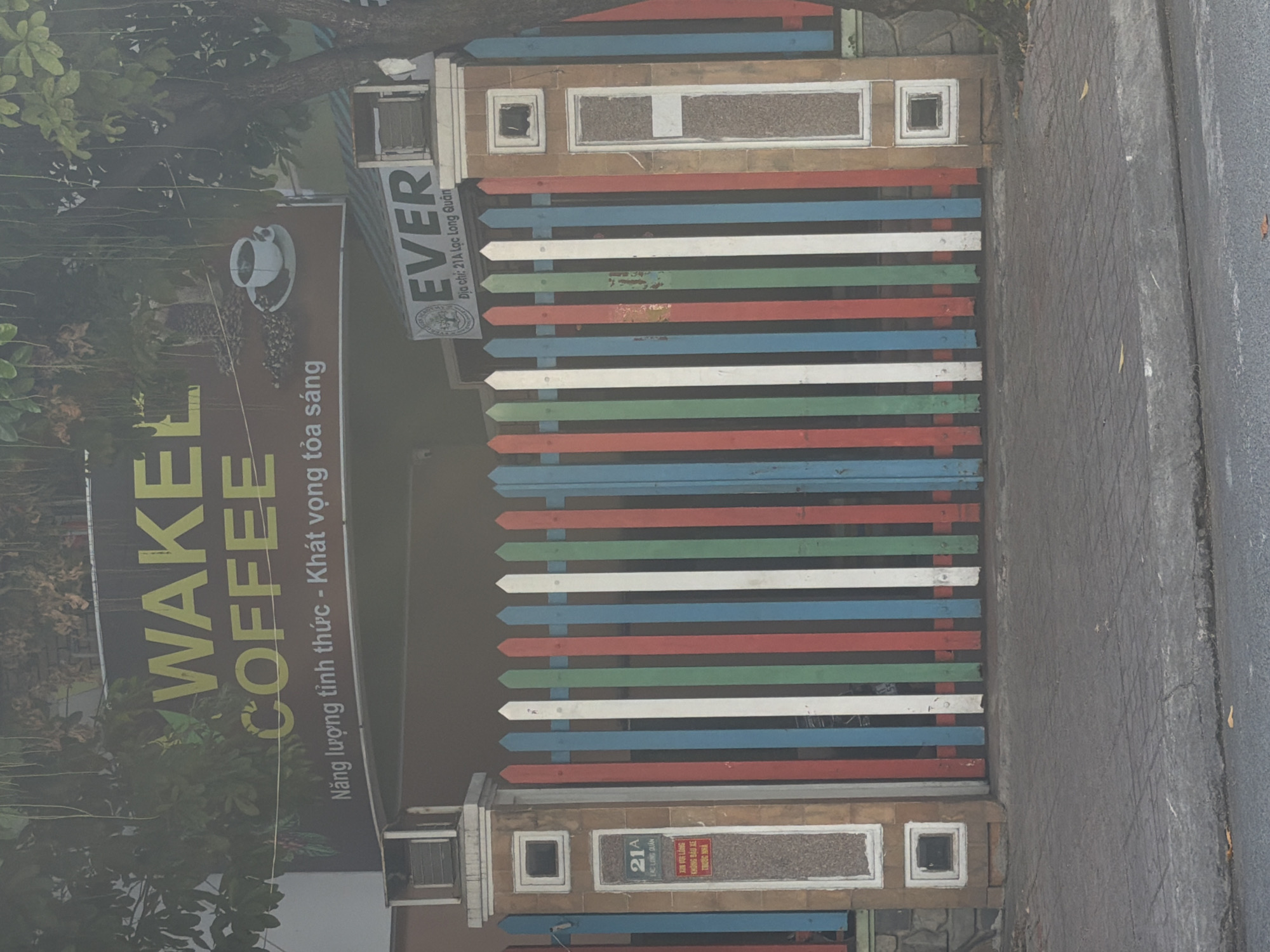 The image shows a colorful wooden gate in front of a building. The gate has vertical slats painted in red, white, green, and blue. On the left side of the gate, a stone pillar has a sign with "21A LẠC LONG QUÂN" written on it. Above the gate, a sign reads "WAKEL COFFEE" in large yellow letters, with smaller Vietnamese text below it. To the right of the "WAKEL COFFEE" sign, another sign displays "EVER" in large letters. The building appears to be a commercial establishment, possibly a cafe. The surrounding environment is somewhat obscured by foliage, suggesting an outdoor setting, likely a street in Nha Trang Ward, Vietnam, given the address on the pillar. The overall impression is one of a vibrant and welcoming entrance.
