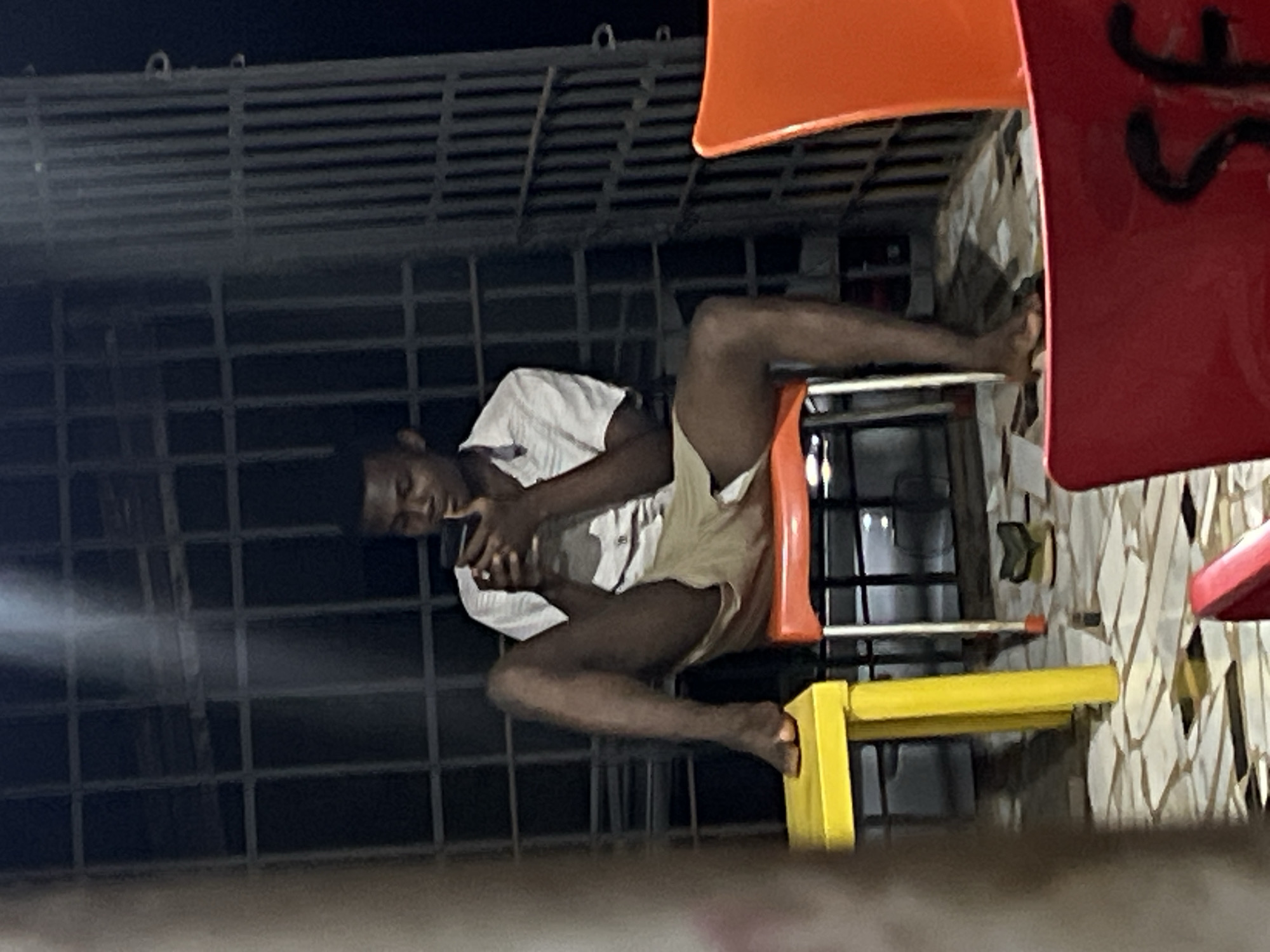 The image depicts a young Black male sitting on a chair in what appears to be an outdoor seating area, possibly at a roadside eatery or bar in Nkpor, Nigeria. He is wearing a short-sleeved collared shirt and khaki shorts and is looking down at his mobile phone. His left leg is extended and his foot is resting on a yellow stool. The background shows a dark metal security grate and some vertical metal bars. In the foreground, parts of red and orange chairs are visible, suggesting multiple seating arrangements. The ground is paved with tiles and there are some scattered papers and a pair of sandals visible. The lighting suggests it is evening or night.