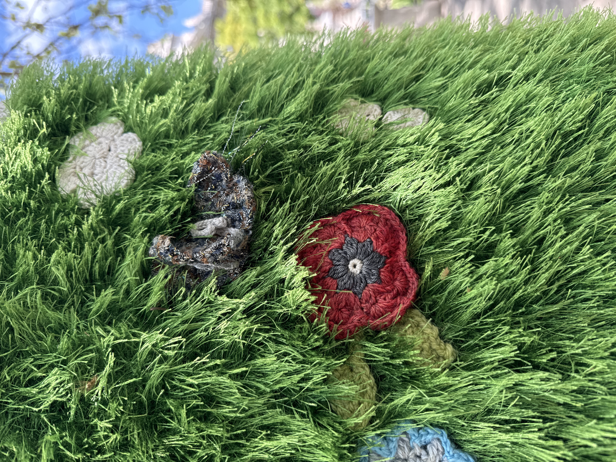 A close-up shot shows a display of knitted objects. The background is soft green strands mimicking grass. On top of the green, there are knitted flowers in colors white and red, and a butterfly in mixed colors. Parts of other knitted objects appear from behind the green strands. The shot is taken during the day with some blue sky visible in the upper left corner. There are buildings in the background, blurred. The scene may be from Budapest, Hungary.