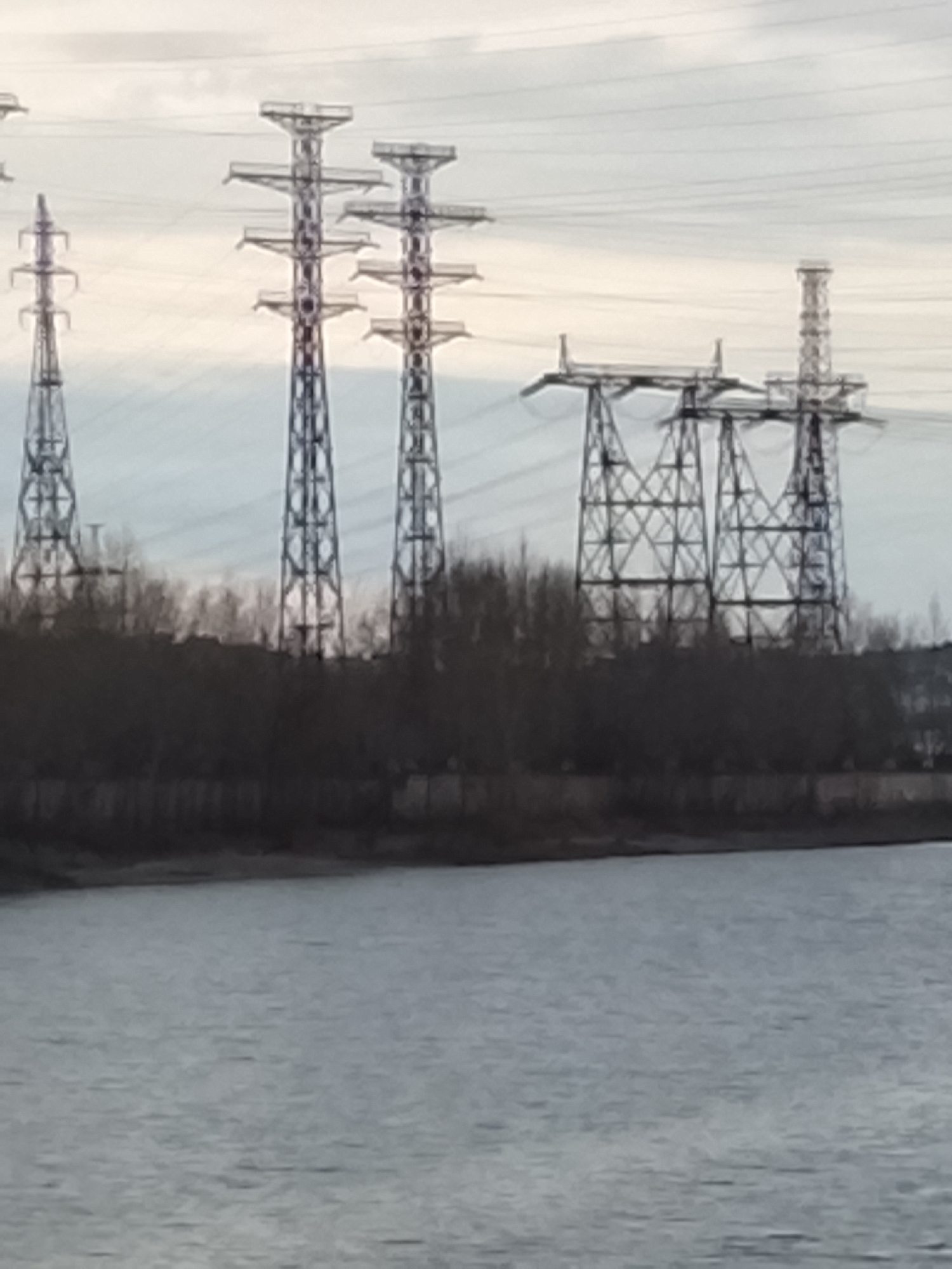 The image depicts a landscape featuring a body of water in the foreground, with a backdrop of land supporting a dense line of bare trees and several electrical transmission towers of varying designs.
The towers, constructed from a latticework of steel, stand prominently against a cloudy sky. Power lines extend between the towers. The scene is urban or industrial, specifically the city of Chaikovsky, Russia, based on the style of the towers that can be found along the Votkinsk reservoir. No people are visible.