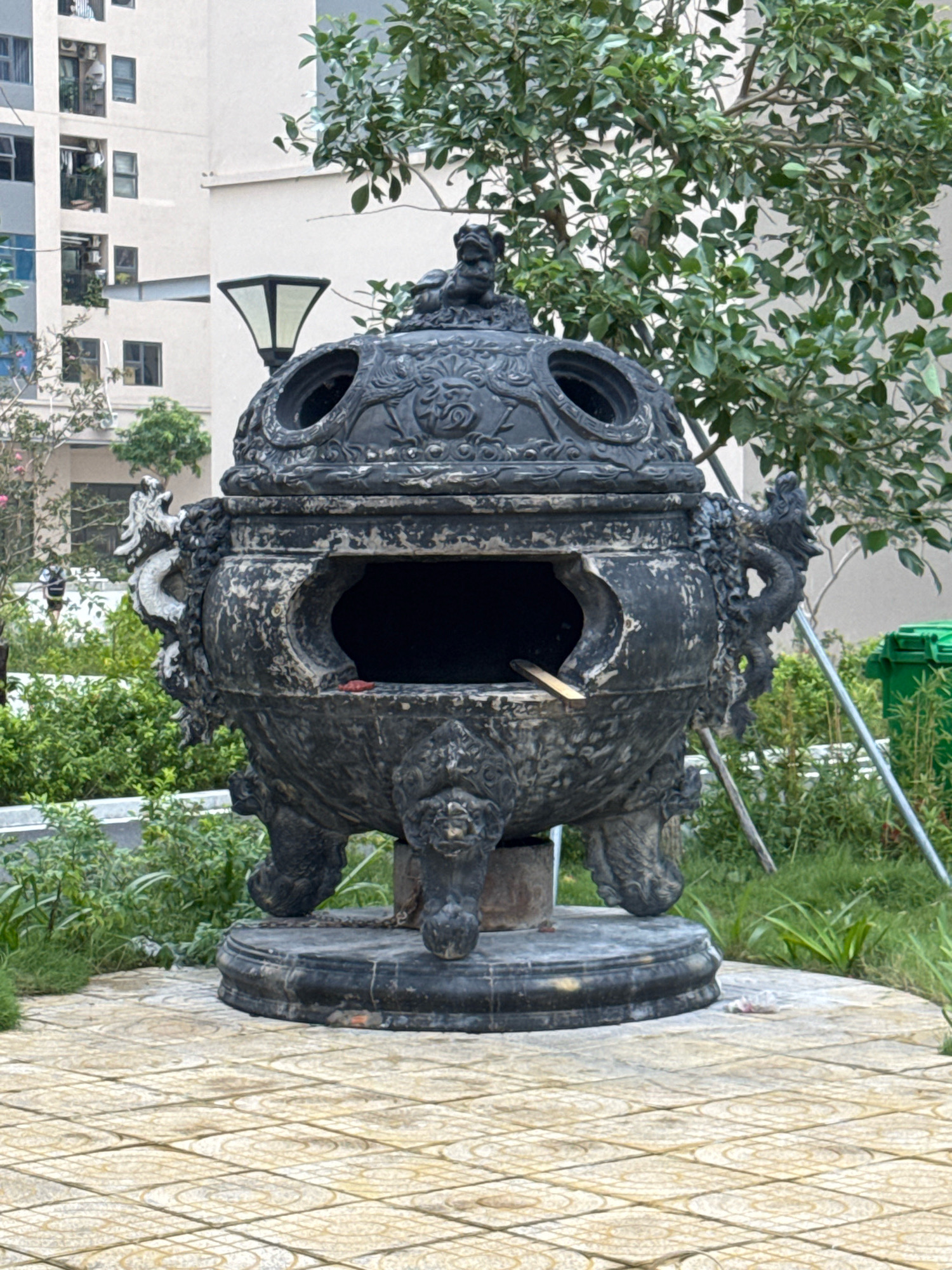 A large, black, ornate incense burner stands in an outdoor courtyard. The burner is intricately decorated with dragon heads on either side and a dragon figure on its lid. It rests on a circular base and has four legs shaped like mythical creatures. The burner has a large opening in its front, revealing a dark interior with a wooden stick protruding. The burner is surrounded by green bushes and trees, and in the background, there is a modern apartment building. The ground is paved with square tiles in a geometric pattern. A green trash bin and a lamppost are also visible in the scene. The lighting suggests it is daytime, possibly late morning or early afternoon.