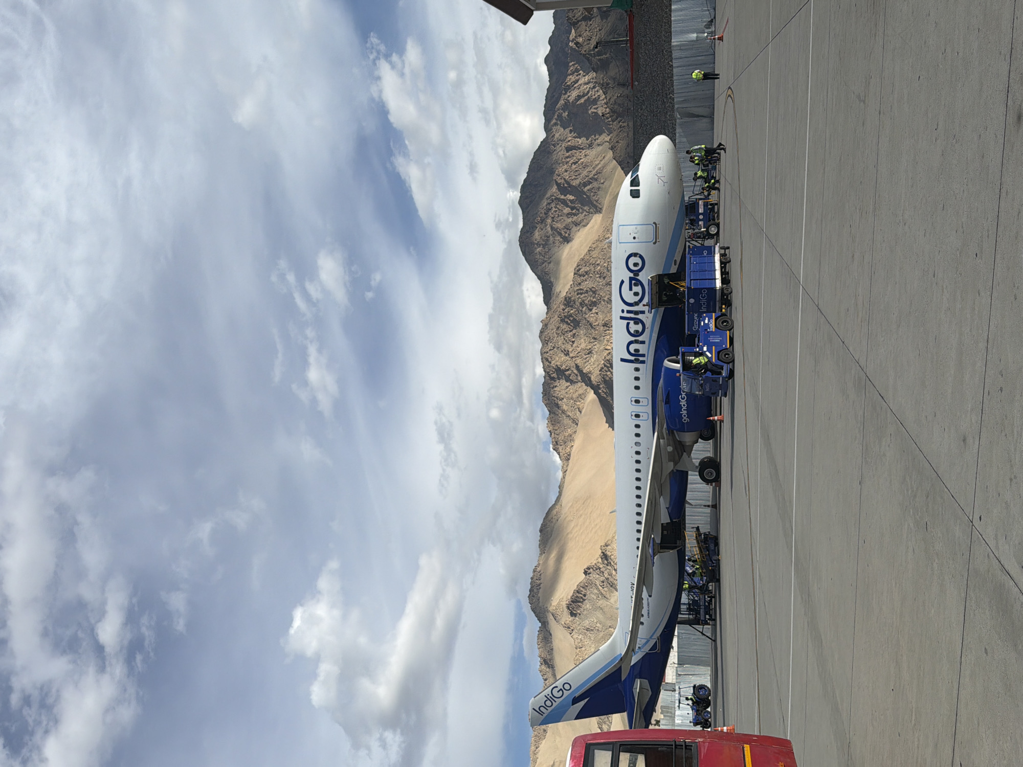 An Indigo Airlines Airbus A320 aircraft is parked on the tarmac at an airport, with a red bus visible to the left. In the background, rugged, barren mountains with patches of snow and vegetation rise under a partially cloudy sky. The aircraft's engines are running, and ground crew members in high-visibility vests are working around it. Luggage carts and other airport vehicles are also present. The scene appears to be at the Leh Kushok Bakula Rimpochee Airport in Spituk, India, given the distinctive mountainous terrain.