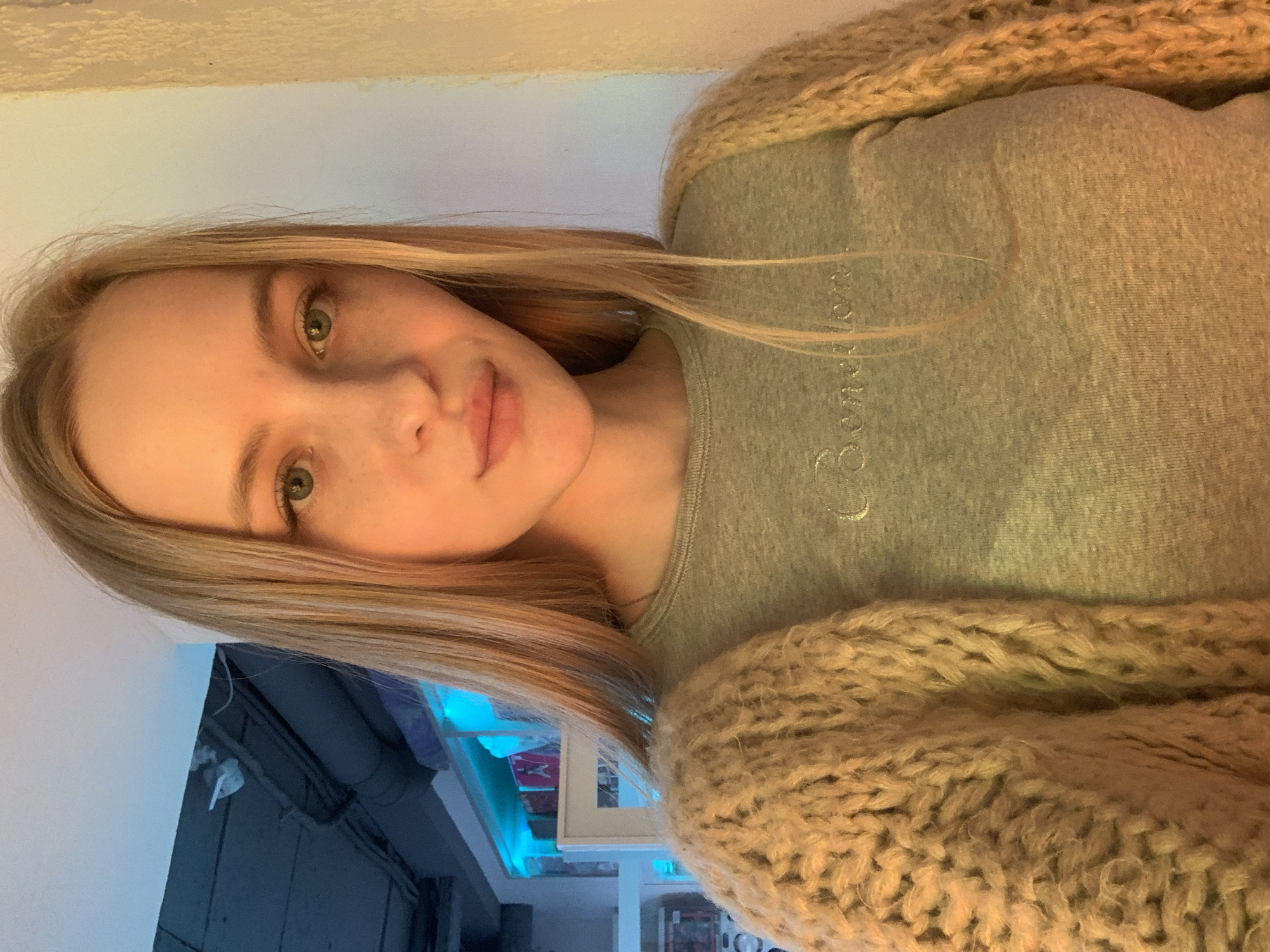The image is a medium shot selfie of a young woman with long blonde hair, freckles, and green eyes. She is wearing a grey long-sleeved shirt with a small embroidered logo and a beige chunky knit cardigan. The background is indistinct, showing what appears to be an indoor space with blue lighting and some industrial-looking pipes on the ceiling, possibly in a loft or studio setting. There are no discernible landmarks or context to suggest a specific location within Kyiv, Ukraine. The woman is looking directly at the camera with a neutral expression.