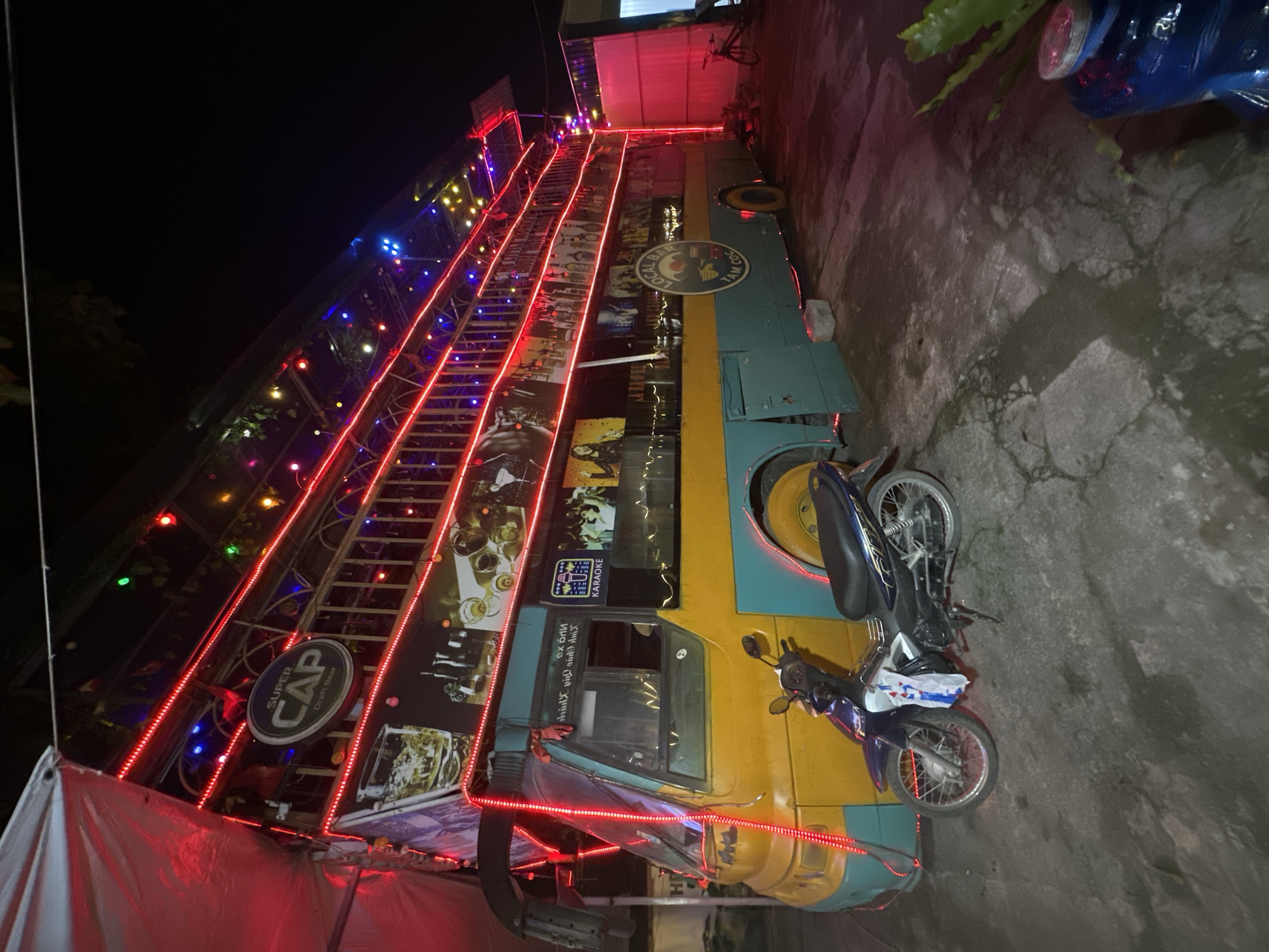 The image captures a unique, double-decker bus converted into a bar or restaurant, situated outdoors at night in Nam Hoa Lư Ward, Vietnam. The bus, painted in vibrant yellow and teal, is adorned with numerous red, blue, and green LED lights that illuminate its exterior, highlighting its multi-tiered structure. A prominent circular sign with the text "SUPER CAP Draft Beer" is visible on the upper level. Various images, including what appear to be drinks and people, are displayed as advertisements or decorations on the side of the bus. A motorcycle is parked in front of the bus. In the background, a bicycle and some greenery are discernible, along with the side of a building featuring pink lighting. The ground is paved with concrete that appears wet in places. The overall scene suggests a lively, nighttime entertainment venue.