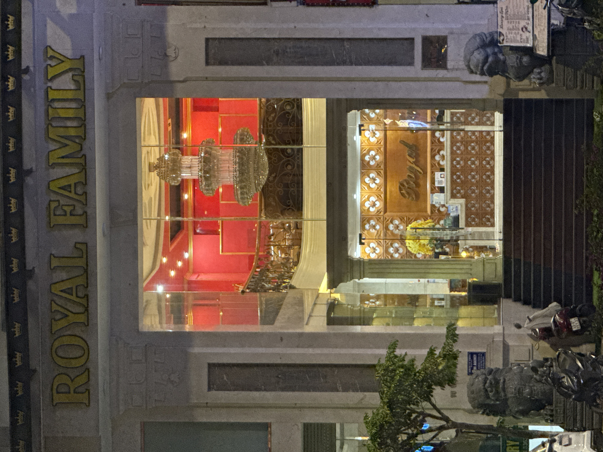 The image shows the exterior of a building in Đà Nẵng, Vietnam, at night. The facade of the building prominently features the name "ROYAL FAMILY" in large, gold lettering running vertically along the left side. Above and below the name, there is a repeated crown motif. The building's architecture appears modern with large windows. Through the windows, ornate interior decorations are visible, including a large chandelier, red walls, and decorative patterns. Motorbikes are parked near the entrance. There are stone sculptures flanking the entrance.