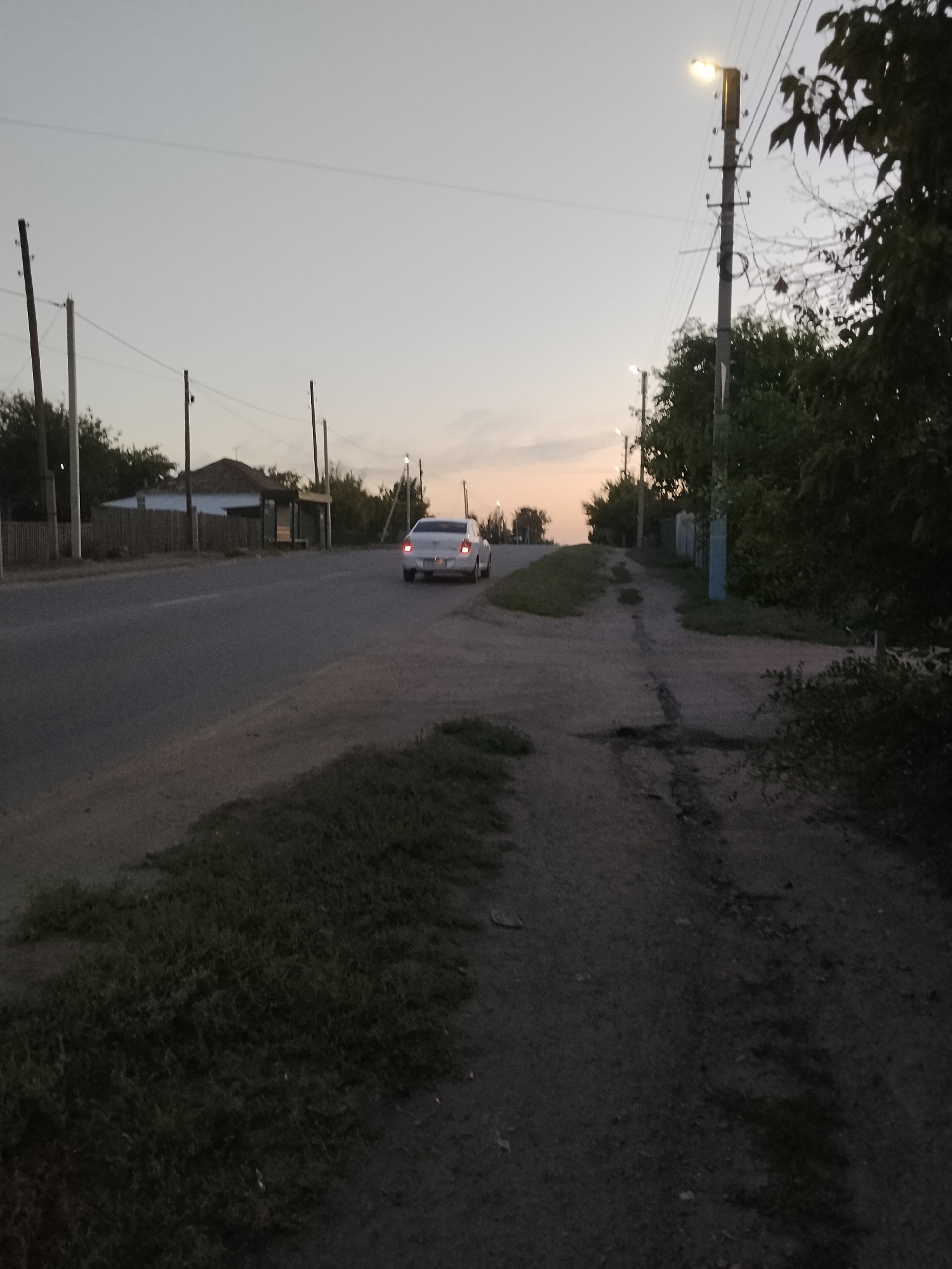 The image captures a street scene in Kokshetau, Kazakhstan, during the twilight hours. The sky is a pale gradient of gray and soft orange, indicating either sunrise or sunset. A paved road runs diagonally from the lower left to the upper right of the frame. On the left side of the road, a low wooden fence borders a property with a house and a small bus stop structure visible behind it. Trees and vegetation line the roadside.
To the right of the road, a dirt path or unpaved shoulder is visible, featuring patches of grass and some darker, potentially damp or worn areas. A prominent utility pole with a lit lamp stands on this side, casting a glow. Further down the road, another similar utility pole is visible.
A white sedan is the only vehicle in motion, seen from the rear as it travels away from the viewer along the paved road. The car's taillights are illuminated. The overall impression is one of a quiet, possibly rural or suburban street in Kokshetau as the day concludes.
