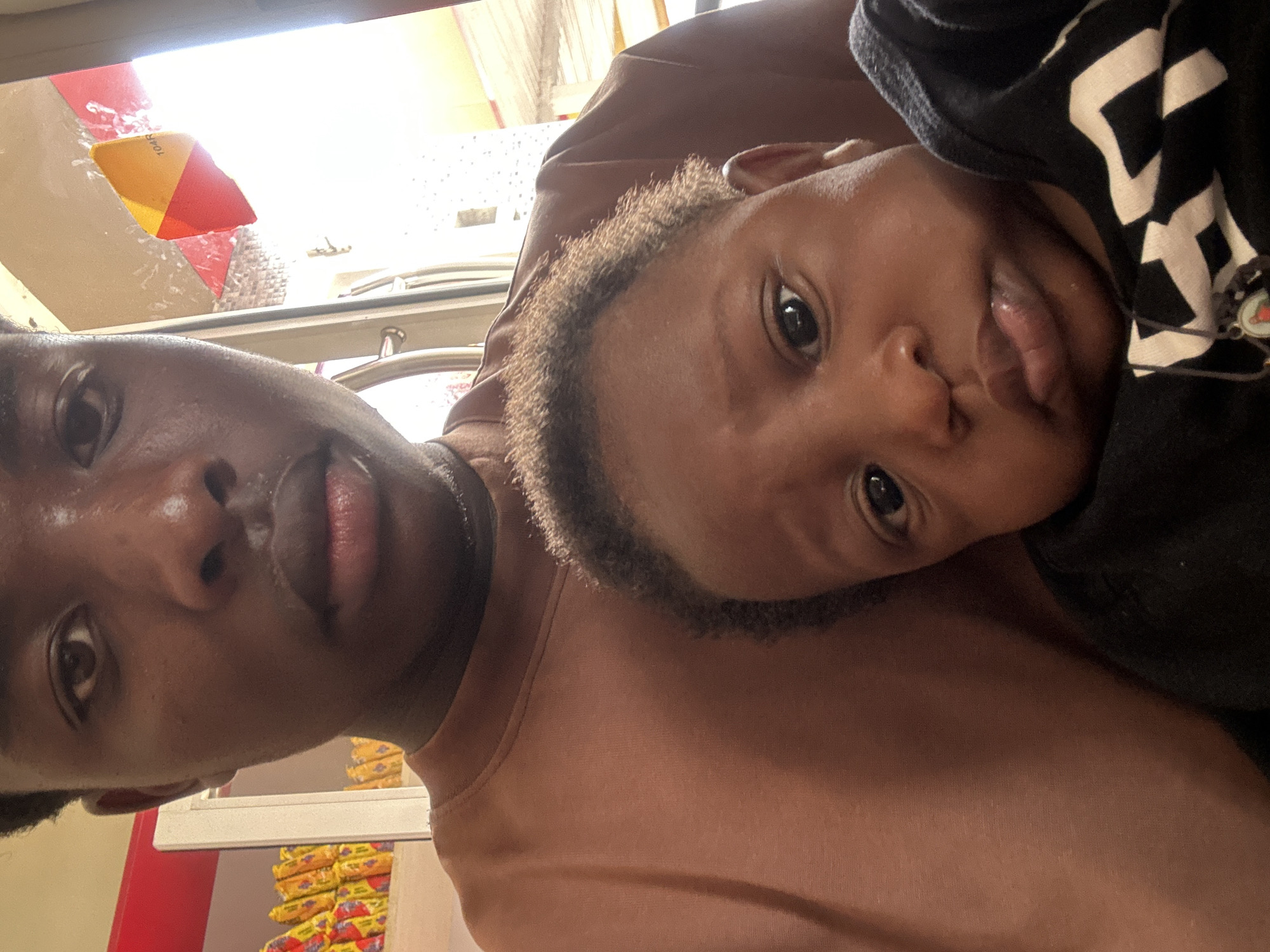 This media file captures a close-up, diagonal selfie of an adult and a child, likely in an indoor commercial setting such as a store or market in Jos, Nigeria. The adult, positioned on the left side of the frame, has dark skin, short dark hair, and is wearing a plain brown or tan shirt. The child, leaning against the adult's shoulder on the right, also has dark skin and short curly hair, dressed in a dark top with white patterns. Both individuals are looking towards the camera with neutral to slightly curious expressions; the child's lips are slightly parted. A small, dark bracelet or charm is visible near the child's shoulder.
The background is brightly lit, with a prominent light source visible in the upper left, possibly a window or overhead lighting. A red and yellow triangular object, possibly a price tag or product label, is attached to a surface in the upper left background and displays the text "104R". Below and to the right of the subjects, numerous stacks of yellow-packaged items with red and purple details are arranged on shelves or a display, characteristic of a retail environment. The image conveys a moment of familial connection or affection between the adult and the child.