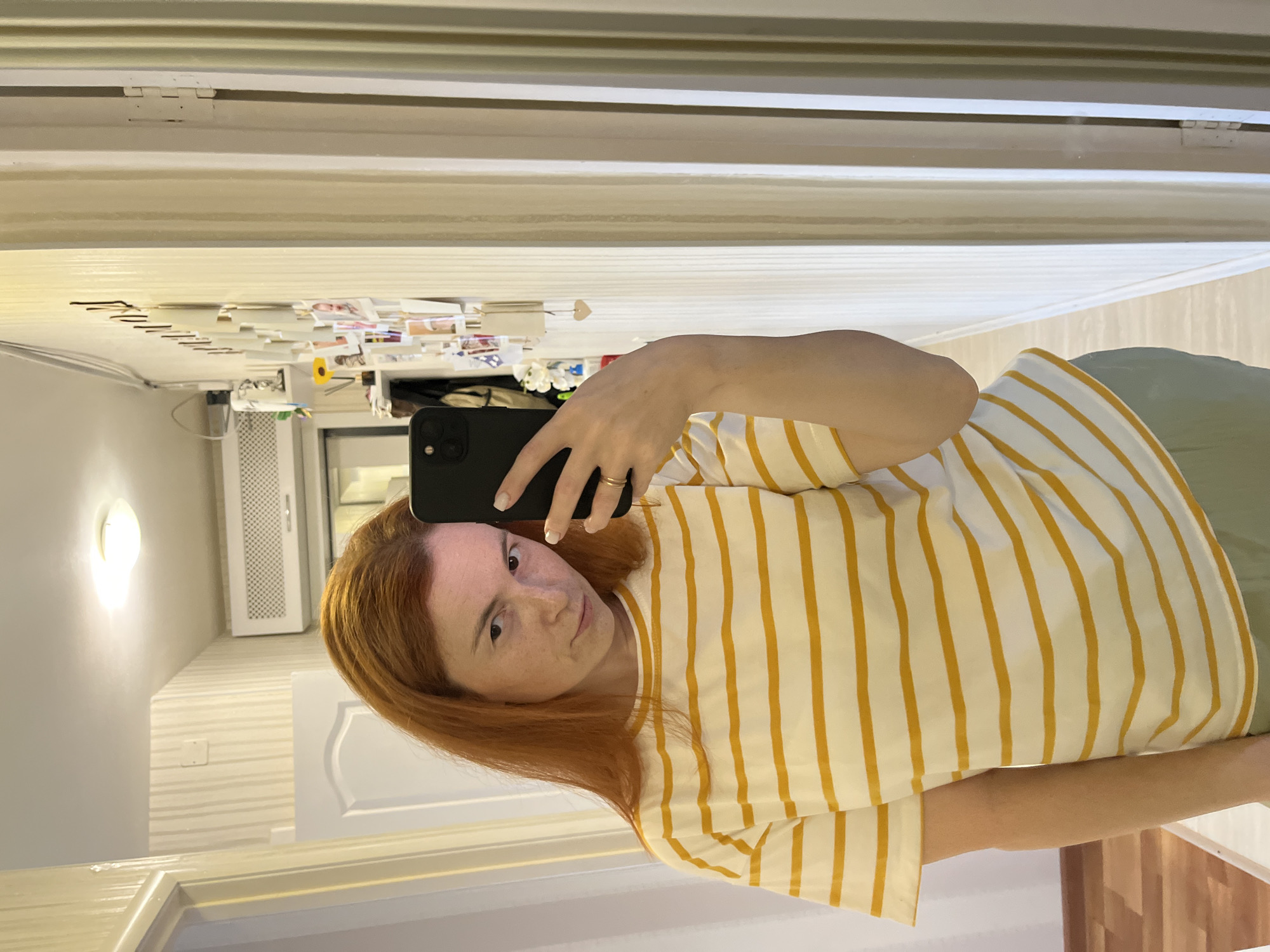 A young woman with long, auburn hair and freckles takes a selfie in a hallway. She is wearing a yellow and white striped t-shirt and light green pants. She is holding a black smartphone in her right hand and looking at the camera with a neutral expression. Her left hand is resting on her hip. The hallway is lit by a ceiling light and has white walls with vertical paneling. There are several framed photos on the wall to her right, and a doorway to her left. An air conditioner is visible on the wall above the doorway.