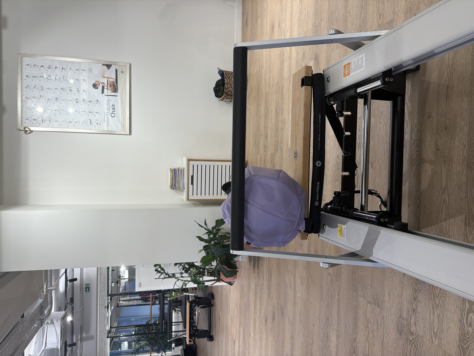 The image depicts a room that appears to be a fitness studio or gym, likely in Boreti, Montenegro. The primary focus is a piece of exercise equipment, possibly a Pilates reformer or stability chair, with a light purple cushion. The equipment has metal and black components, and there's visible branding text that reads "STOTT PILATES complete Stability Chair" and "merrithew. INTELLIGENT EXERCISE, EXCEPTIONAL EQUIPMENT". A caution sticker is also visible on the equipment.
In the background, a framed poster hangs on a white wall, showing illustrations of various exercises on a stability chair. To the left of the equipment, a woven basket hangs on the wall, containing some items. Further to the left, a white cabinet with slatted doors is visible. In the foreground and to the left, a lush green plant in a pot adds a touch of nature to the scene.
The room has light-colored wooden flooring and white walls, contributing to a clean and modern aesthetic. The lighting suggests it is daytime, possibly indoors, as there are no clear external weather cues. The overall impression is of a well-equipped and tidy fitness space, ready for use.