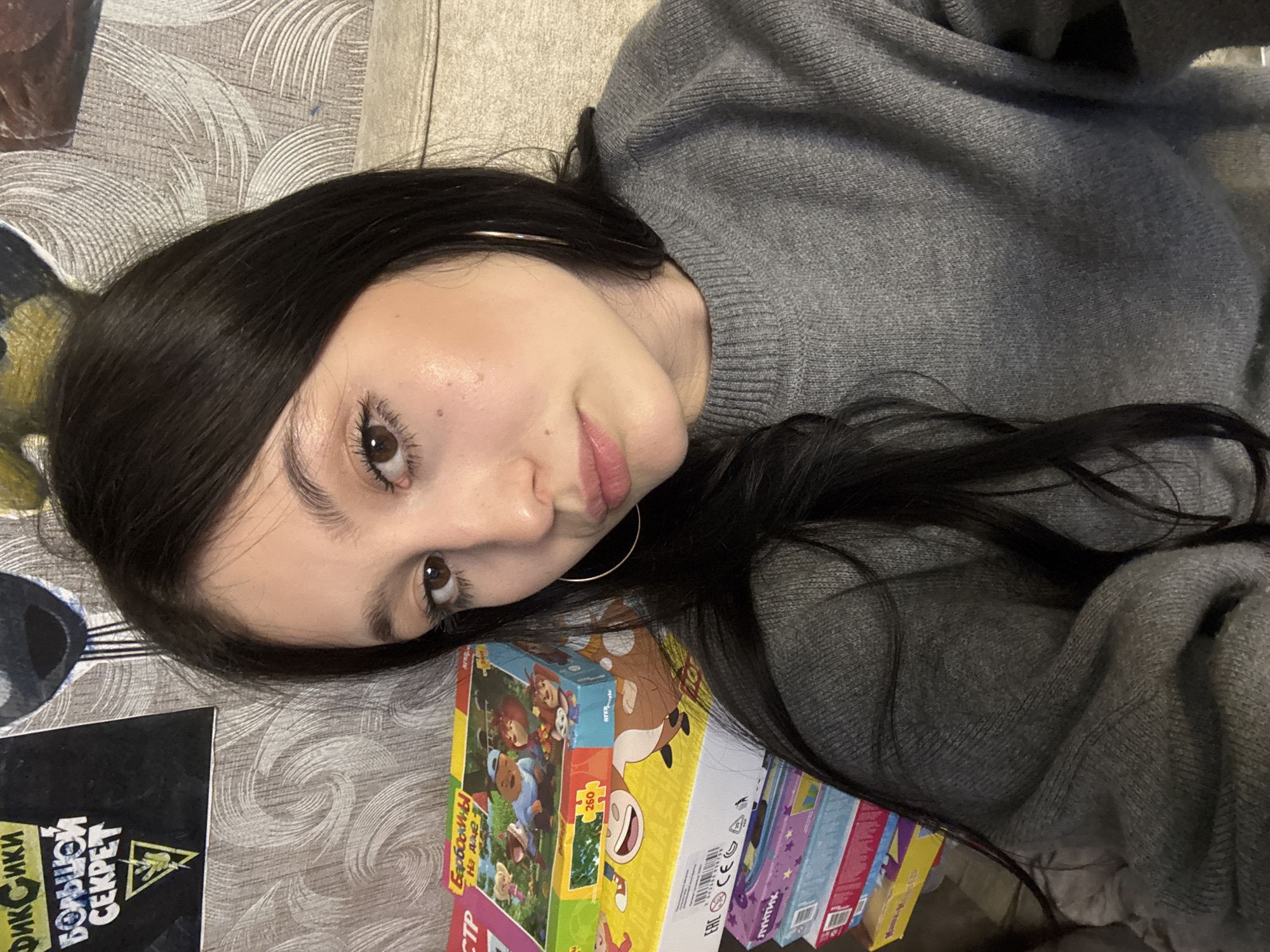 A close-up, high-angle indoor selfie captures a young female subject in the city of Фоки, Russia. The individual has long, dark brown hair, dark brown eyes, and fair skin, with visible makeup on her eyes and lips. She is wearing a grey knitted crew-neck sweater and a gold-colored hoop earring on her left ear, looking directly at the camera with a neutral expression and slightly parted lips. To her lower left, a stack of colorful cardboard boxes, identified as children's puzzles or games, is present. The topmost visible box features characters from "Барбоскины" (Barboskiny) and the number "260," while lower boxes show a cartoon cow and partially visible "ЛУНТИК" (Luntik) branding. On the wall behind and to the upper left, a black poster with white and yellow text reads "ФИКСИКИ БОЛЬШОЙ СЕКРЕТ" (FIXIKI THE BIG SECRET) above a triangular symbol. The wall itself is covered in greyish-brown wallpaper with a swirling pattern. To the right of the subject, a light brown, textured upholstered surface is partially visible, consistent with a couch or bed.