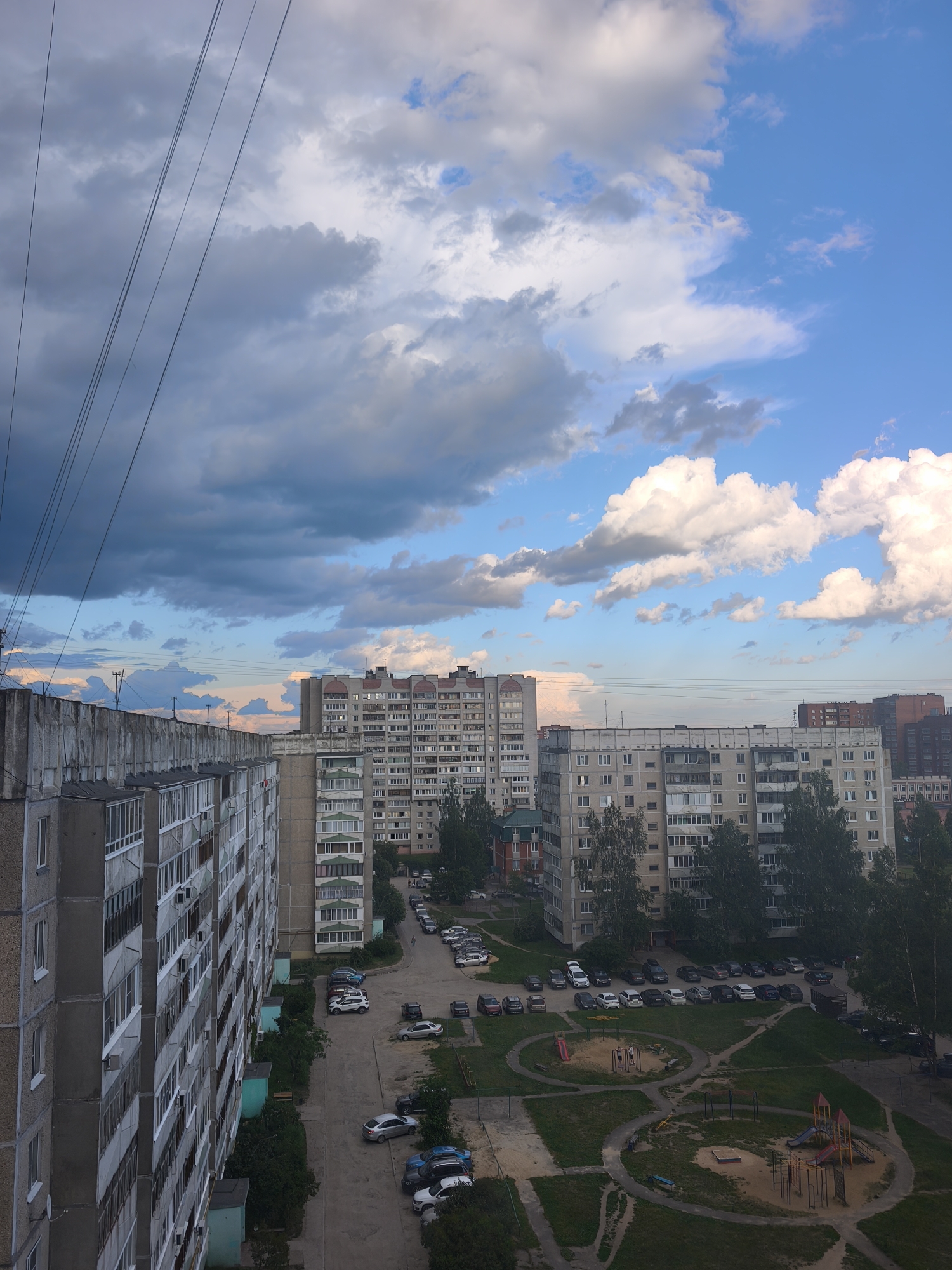 This is a high-angle, full shot of a residential area in Yoshkar-Ola, Russia, under a dynamic sky. The foreground features the side of a large, grey, multi-story apartment building with numerous balconies. To the right, another similar apartment building of lighter color is visible, extending further into the frame. Beyond these buildings, more apartment blocks are seen, varying in height and architectural style, with some displaying visible windows and balconies. A paved area with parked cars is situated between the buildings, indicating a common urban parking arrangement. A children's playground with slides and swings is visible in the mid-ground, surrounded by green grass and trees. Power lines crisscross the upper portion of the image, strung against a sky filled with a mixture of dramatic, dark grey clouds and bright, white cumulus clouds, interspersed with patches of clear blue sky. The overall impression is that of a typical Soviet-era residential development on a cloudy day, with the overcast sky suggesting a recent or approaching weather change. The scene is static and devoid of any visible human action or interaction, focusing on the urban landscape and the atmospheric conditions.