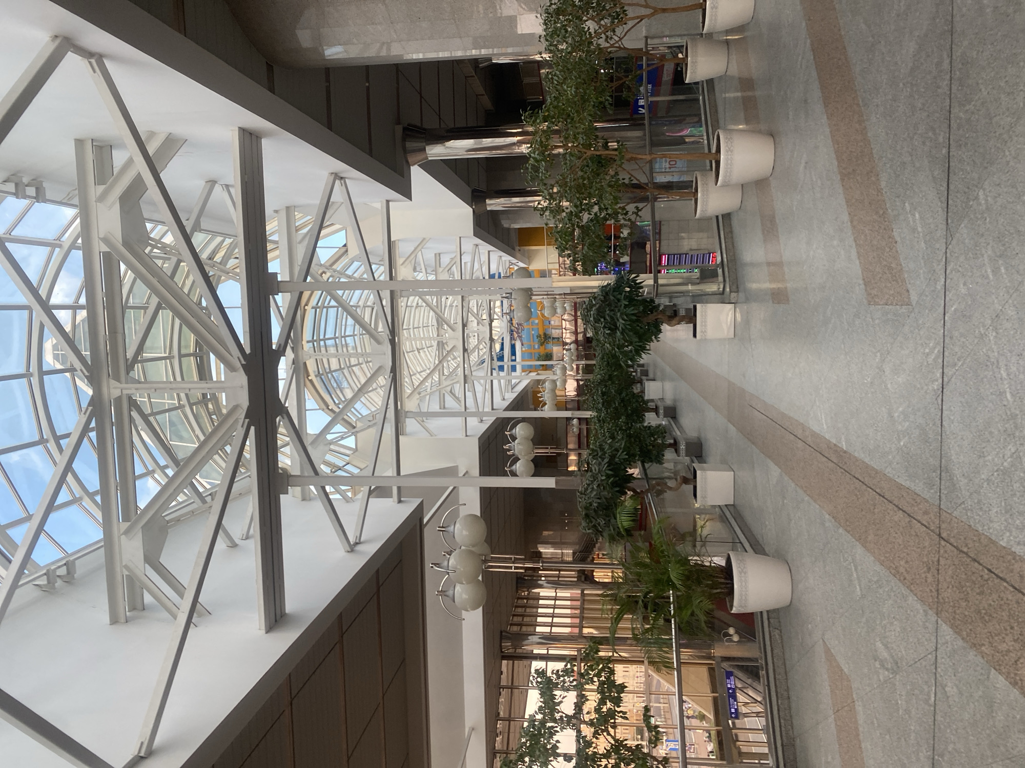 The image depicts the interior of a modern shopping mall or transit hub, likely in Minsk, Belarus, characterized by its expansive glass ceiling and intricate white metal framework. The space is filled with potted plants, including ficus trees and ferns, strategically placed along a polished marble floor. The floor has a patterned design with light grey and pinkish-brown rectangular inserts. Several clusters of spherical white lights hang from poles, illuminating the walkway. Large, polished metal columns support sections of the ceiling. The background shows glimpses of shops and escalators through glass partitions and railings. The overall atmosphere is bright and airy, with natural light filtering through the transparent roof. There are no people visible in the frame.