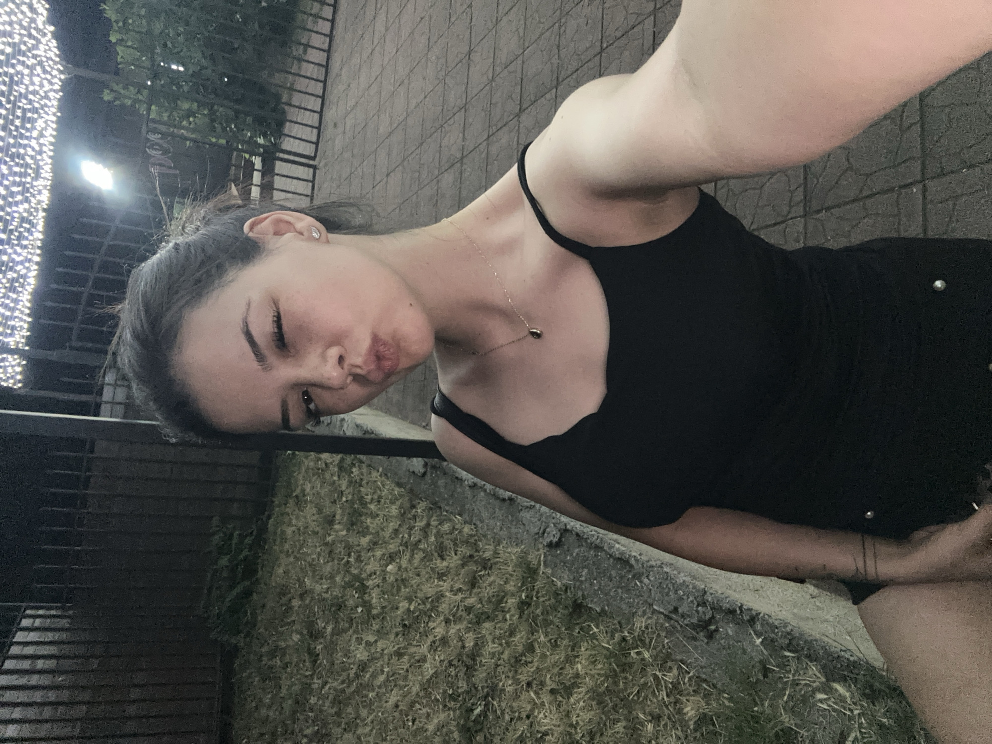 A young woman with dark hair pulled back, wearing a black tank top and a gold necklace, is taking a selfie outdoors at night. She is winking and puckering her lips. Behind her, a black metal fence encloses an area with dry grass, a paved walkway, and a canopy adorned with string lights. The setting appears to be a public space in the city of Sliven, Bulgaria.