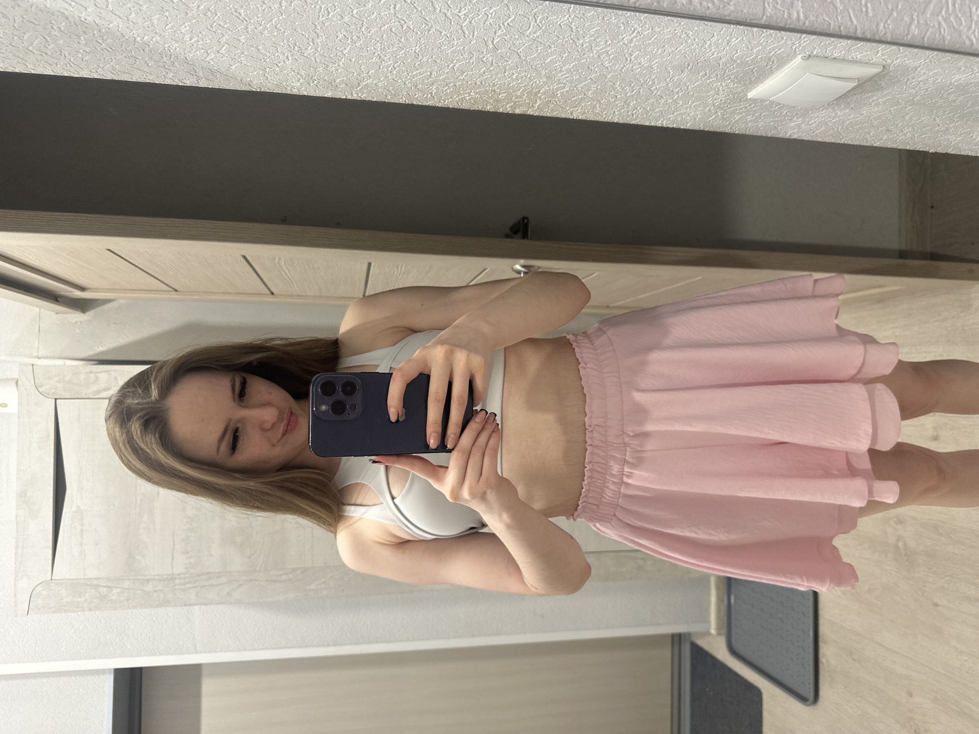 A young woman is taking a selfie in a mirror. She is wearing a white sports bra and a pink skirt. The background is a wall and a doorframe. The location is unknown, Unknown.
