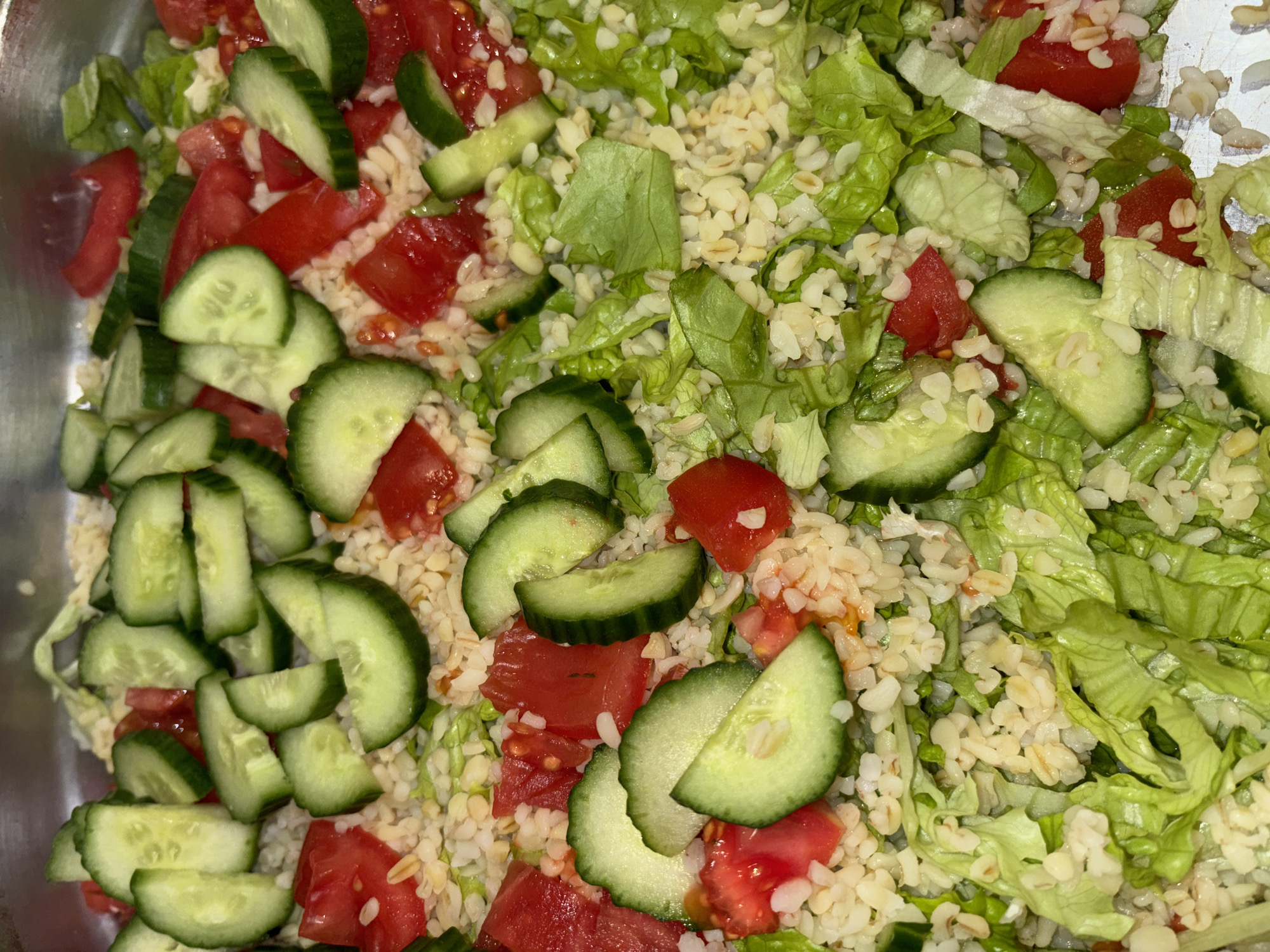 A close-up shot of a salad with tomatoes, cucumbers, lettuce, and bulgur wheat. The salad is contained in a metal bowl or container. The cucumbers are sliced into half-moons and the tomatoes are diced. The lettuce is coarsely chopped. The bulgur wheat forms the base of the salad. The background is blurred and out of focus. The location of the photo is not discernible.