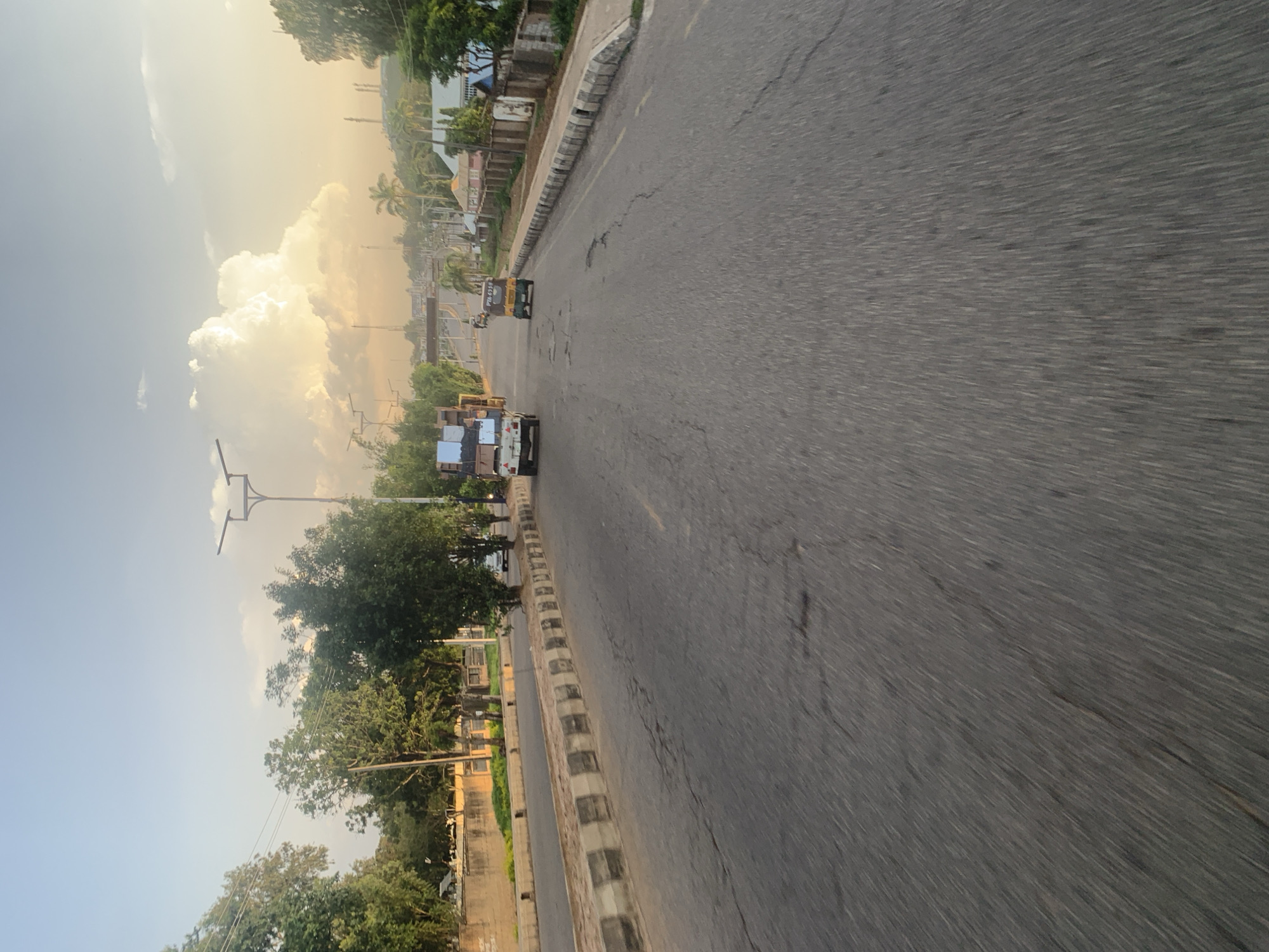 The image depicts a street scene in Jos, Nigeria, on a clear day with a bright sky and scattered clouds. The perspective is from a moving vehicle, creating a sense of motion blur across the asphalt road. The road itself is cracked and worn in places, with a distinct median bordered by concrete blocks.
On the right side of the road, a low concrete wall with regularly spaced vertical concrete posts separates the road from a grassy area and some buildings. These buildings appear to be residential or commercial, with some featuring colorful facades and fenced perimeters. Mature trees line the right side of the road, casting shadows.
On the left side of the road, a similar concrete wall and grassy area are visible, with more trees and some structures in the background. The street is moderately trafficked, with a few vehicles present. In the middle ground, a white pickup truck loaded with items is seen, and to its right, a yellow three-wheeled auto-rickshaw is traveling in the same direction. Further in the distance, an overpass or bridge can be seen, with more vehicles and what appear to be streetlights and utility poles. The overall environment suggests an urban or semi-urban setting within Jos.